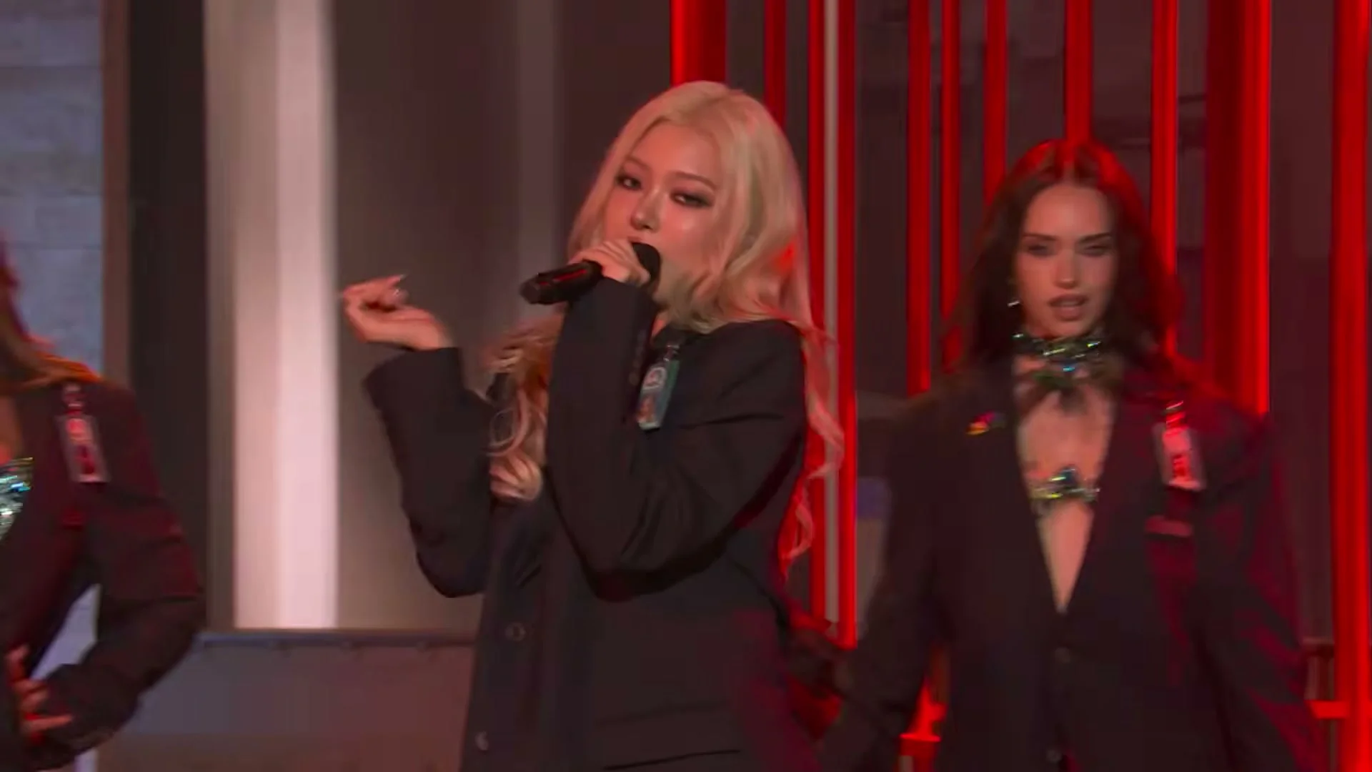 A KATSEYE singer holds a microphone while another member dances behind her during Animal on SNL.