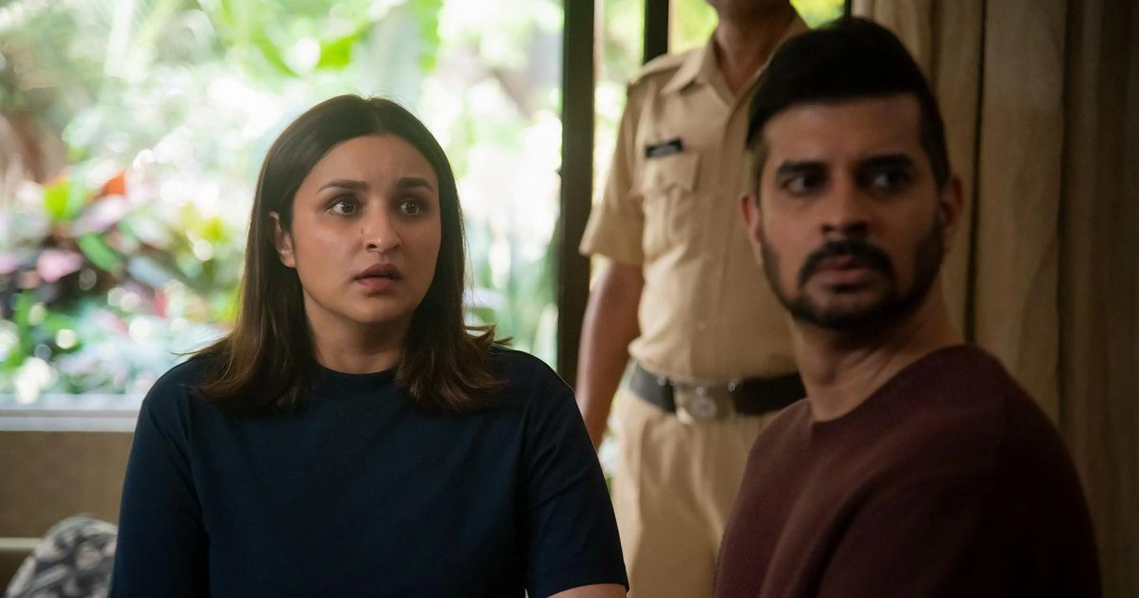 Parineeti Chopra as Anvita and Tahir Raj Bhasin as Alok look towards someone in a police station, with a uniformed officer behind them.