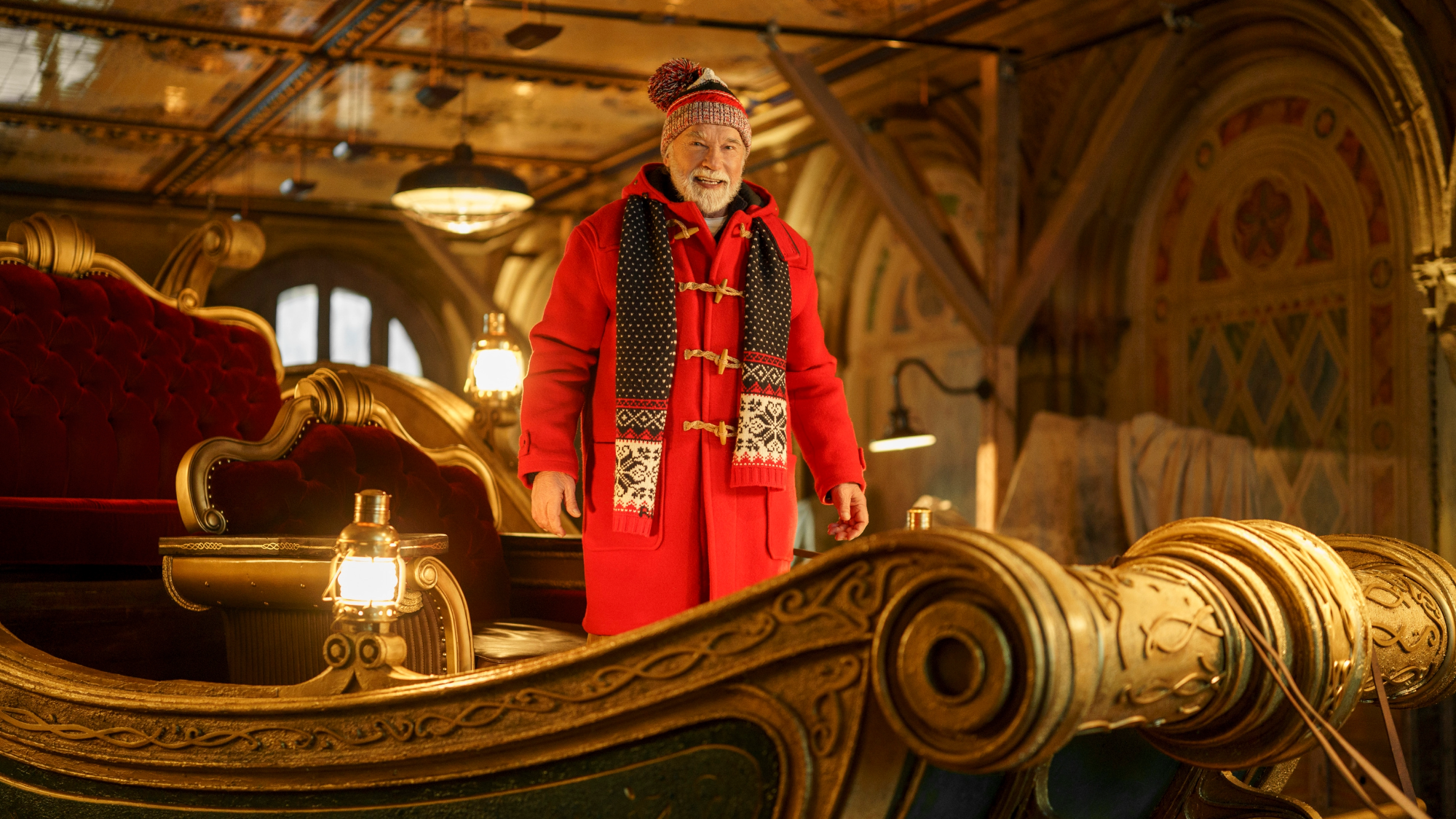 Arnold Schwarzenegger as Santa, wearing a red coat and knit hat on an ornate sleigh.
