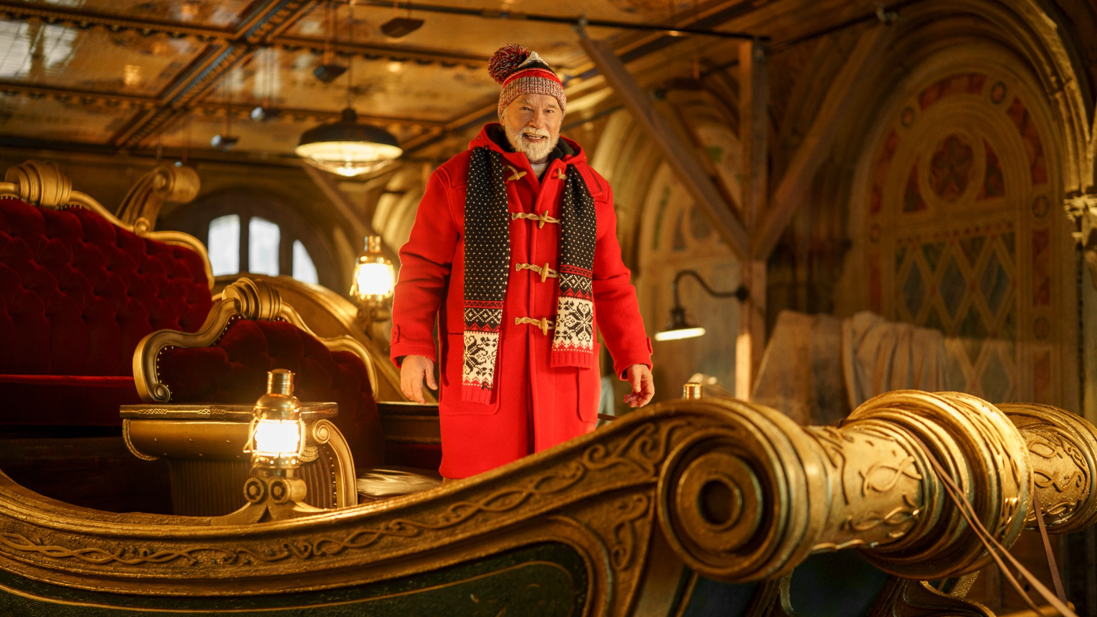 Arnold Schwarzenegger as Santa, wearing a red coat and knit hat on an ornate sleigh.