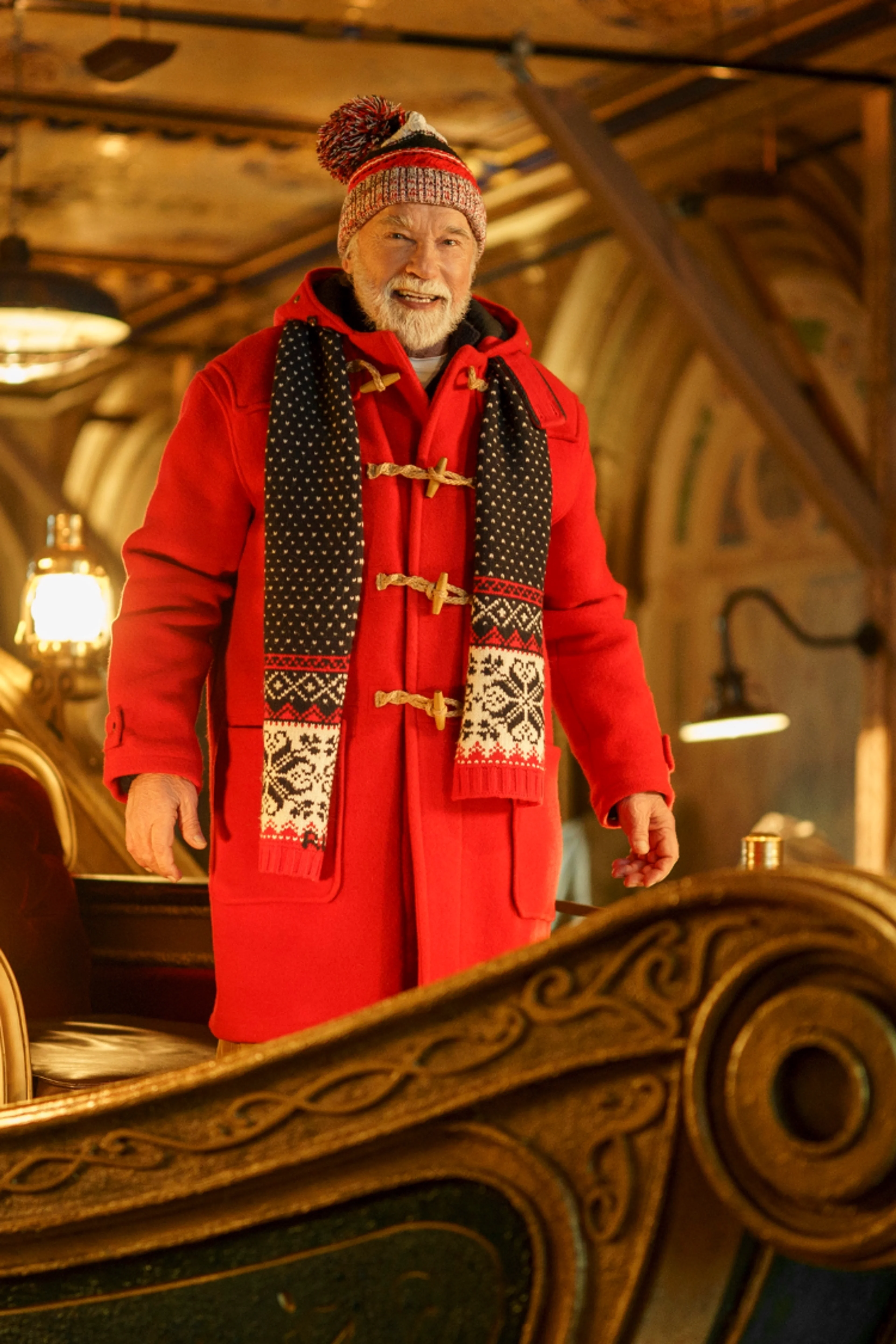 Arnold Schwarzenegger as Santa, wearing a red coat and knit hat on an ornate sleigh.