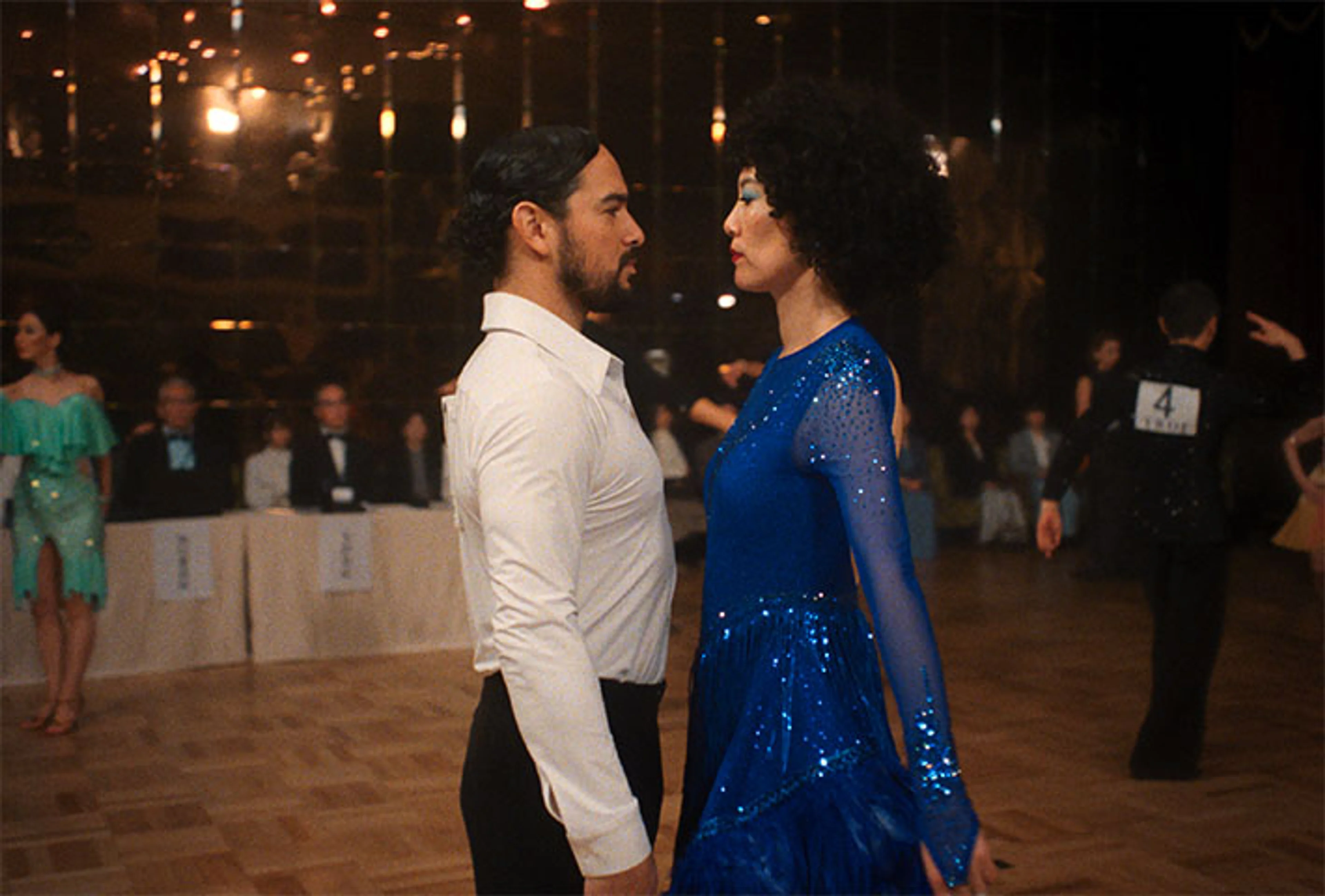 Rinko Kikuchi in a blue dance dress faces a partner on the ballroom floor.