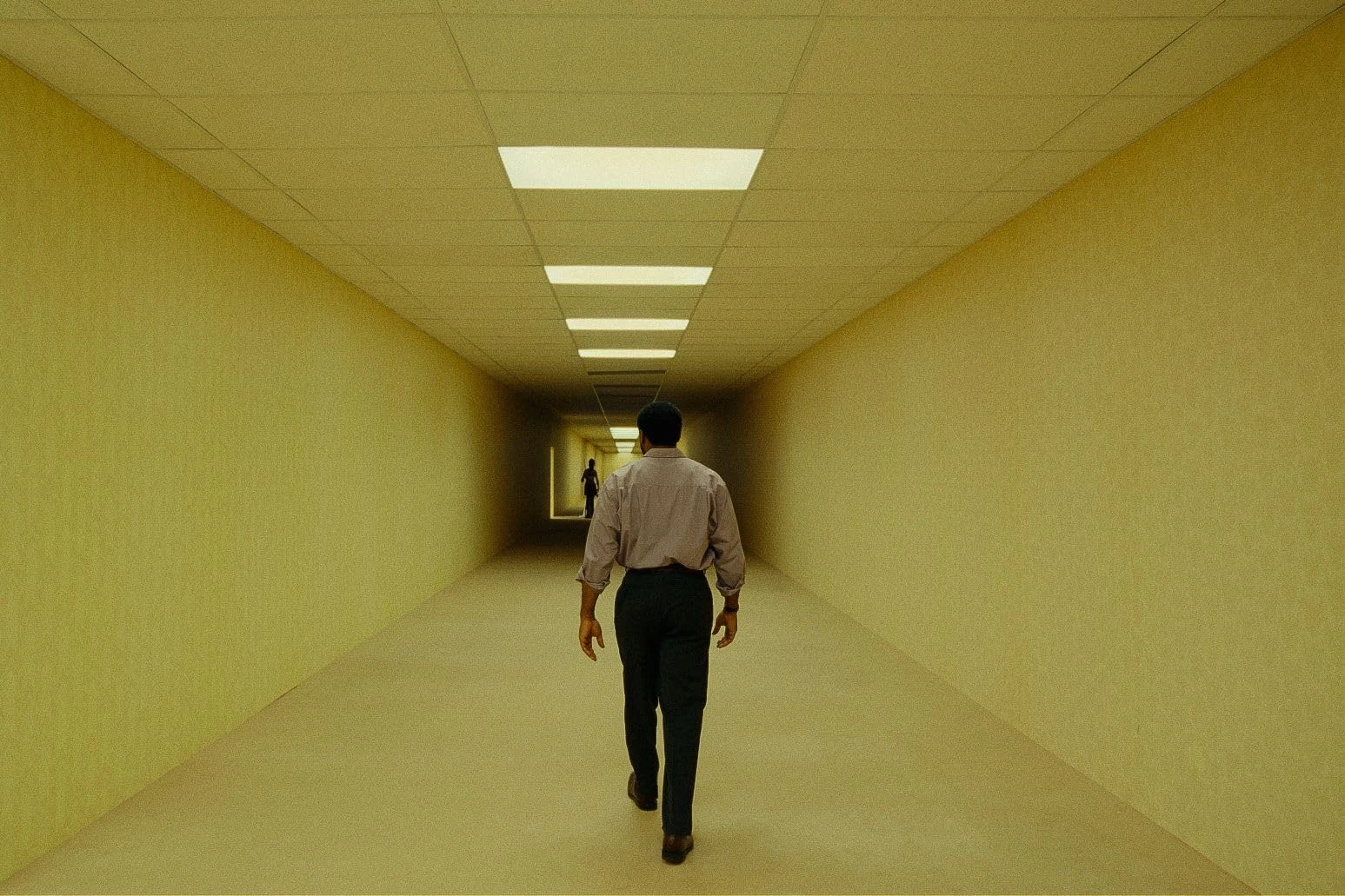 A man walks toward a distant figure in a long yellow hallway lit by fluorescent ceiling panels.