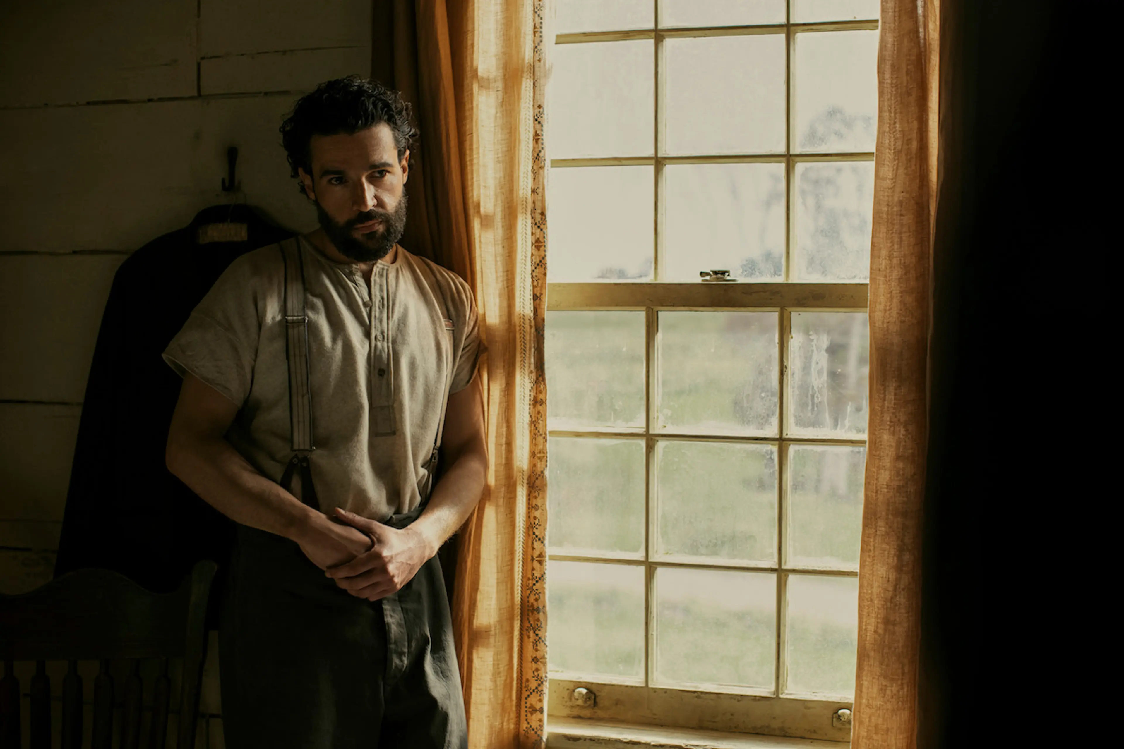 A bearded man in period clothing stands beside a sunlit farmhouse window in East of Eden.