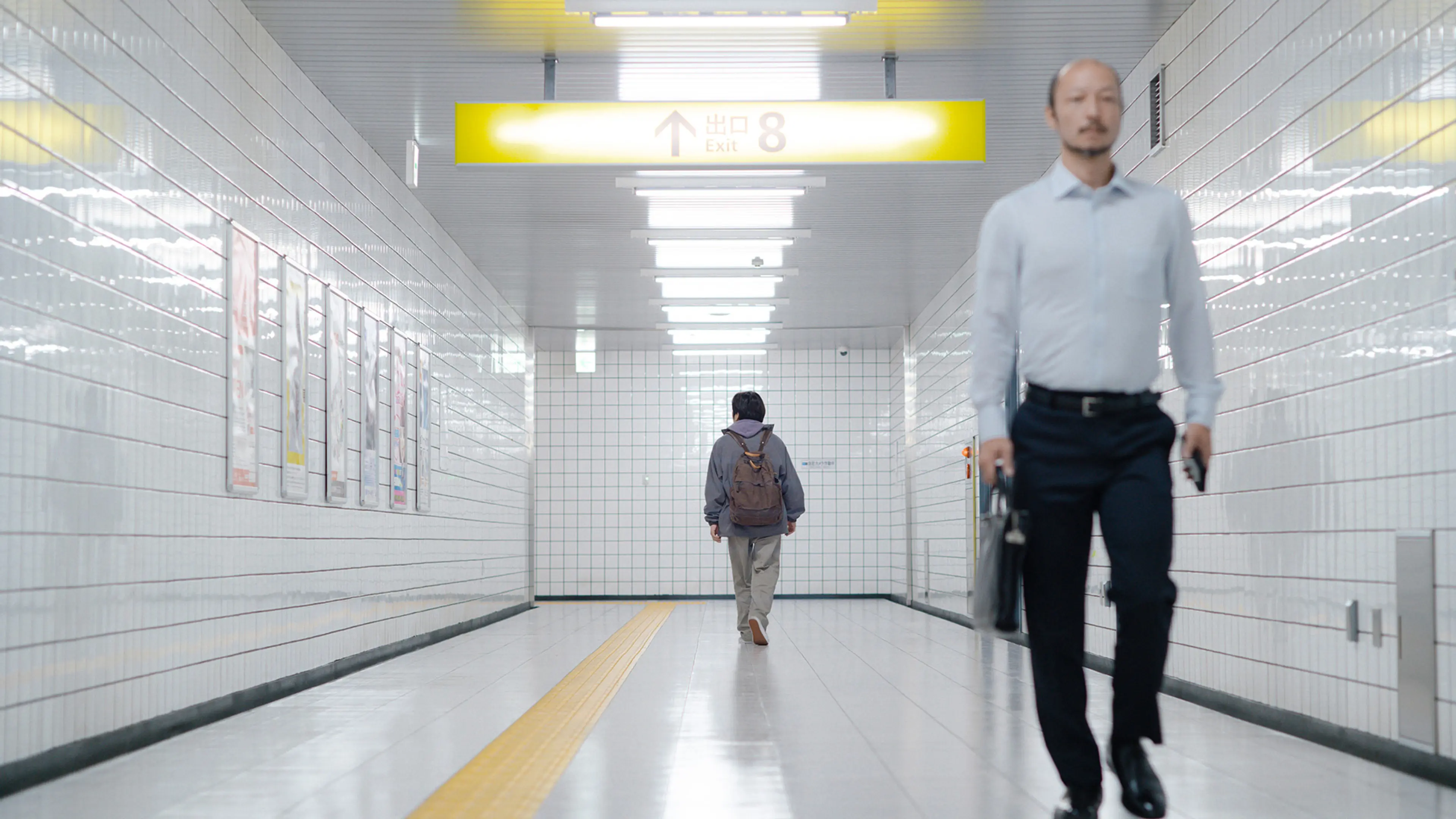 A man in a white shirt fills the foreground while a backpack-wearing traveler walks away behind him.
