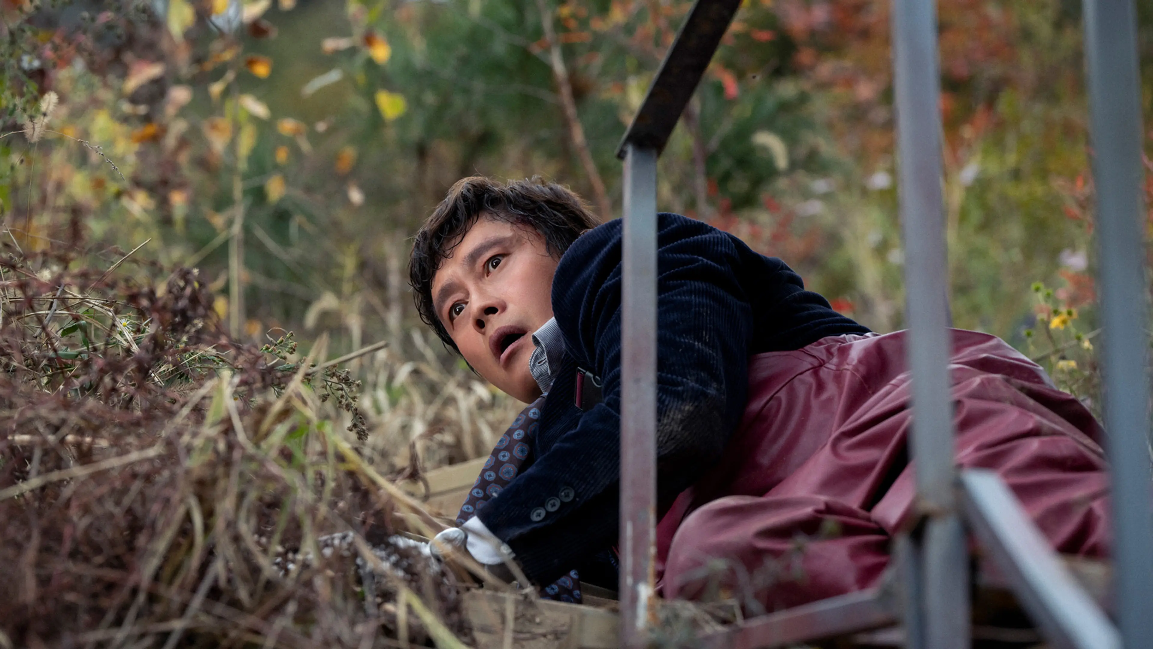 Man-su crouches beside a metal fence in the woods, wearing his red apron.