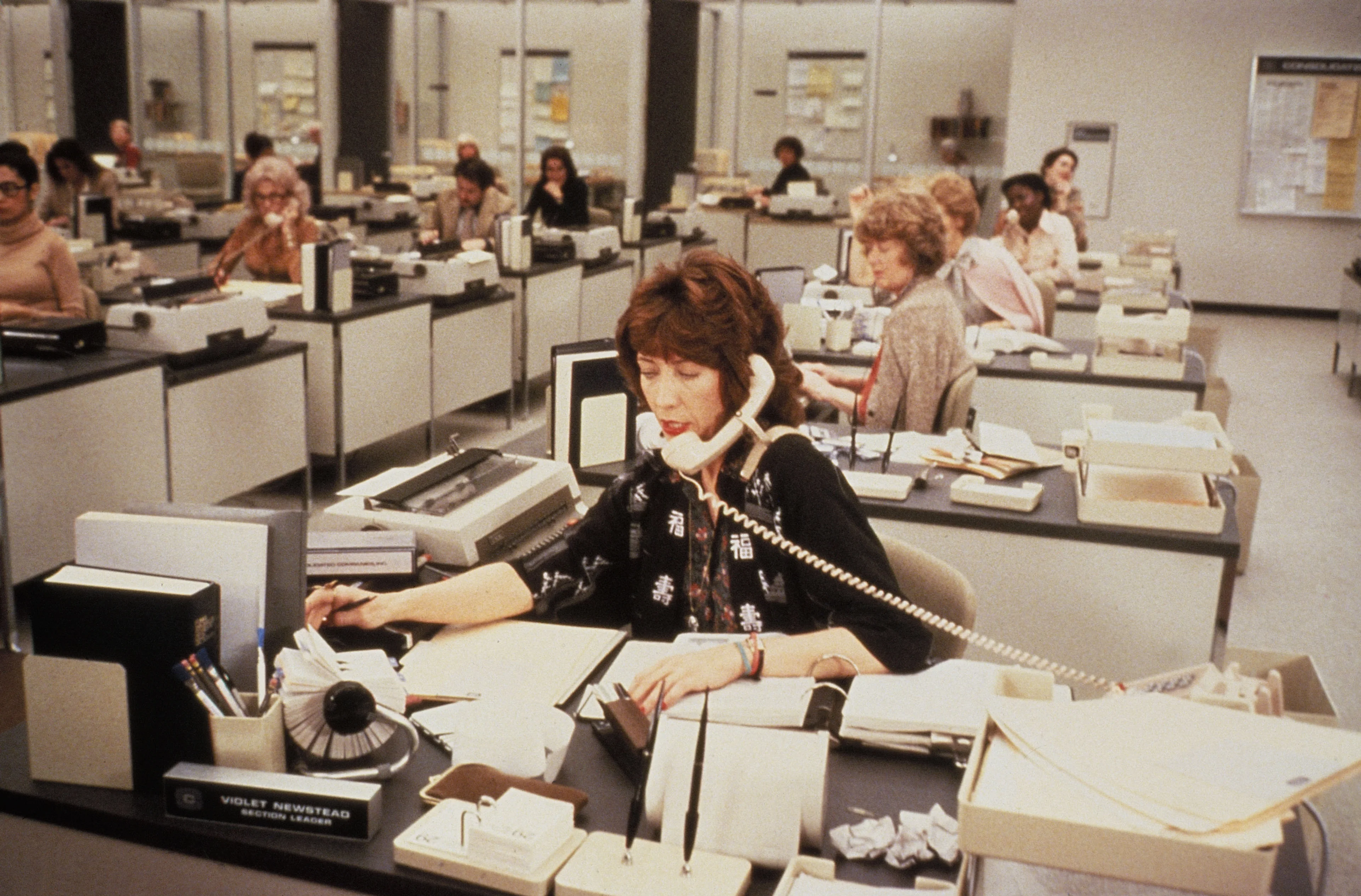 Lily Tomlin at her desk in a busy office scene from 9 to 5.
