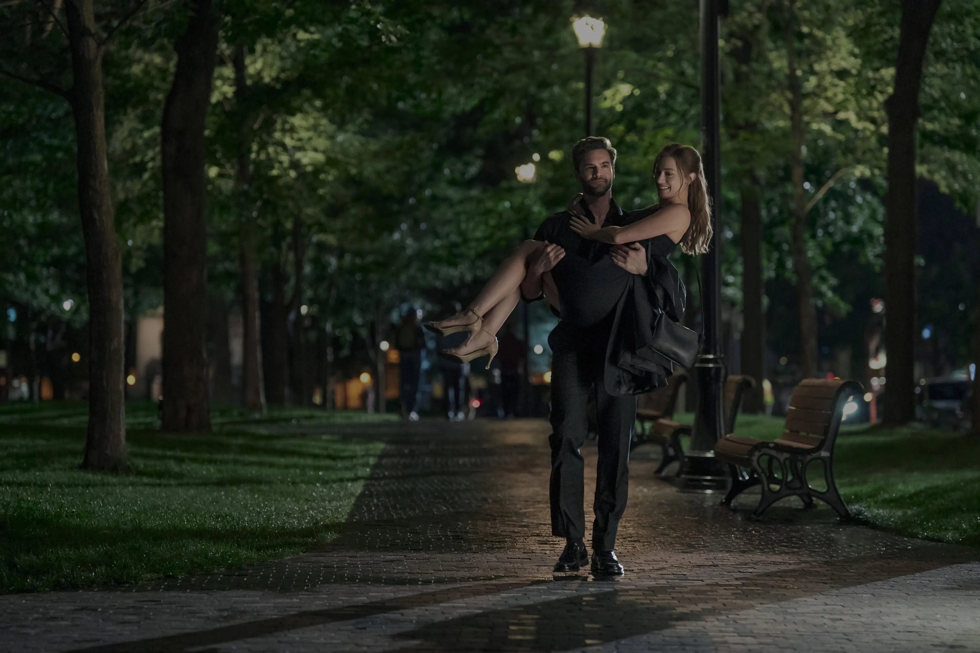 Adam carries Olive along a tree-lined path at night.