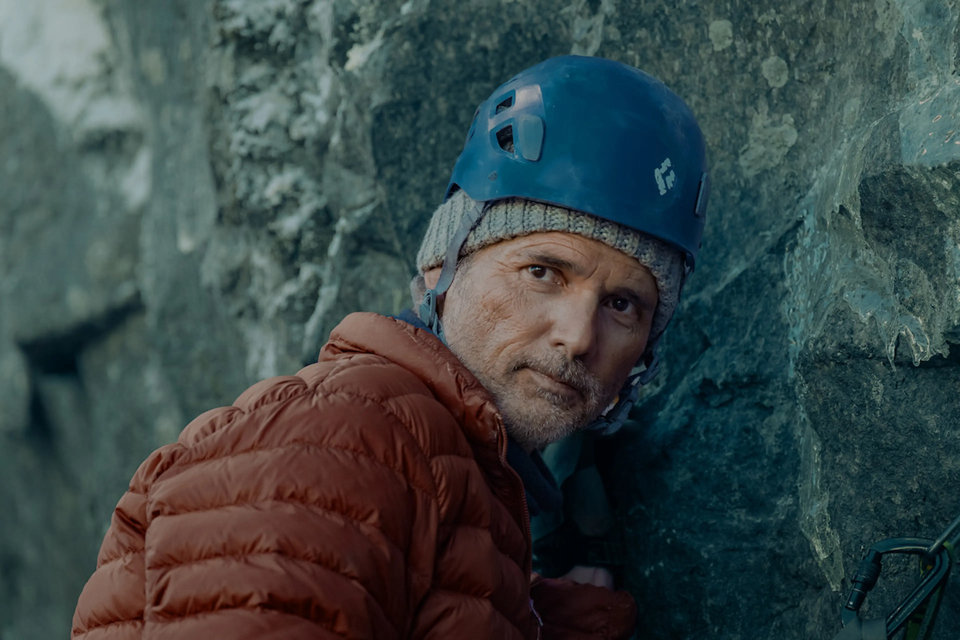 Eric Bana as Tommy wears a blue climbing helmet and an orange jacket beside a cliff.
