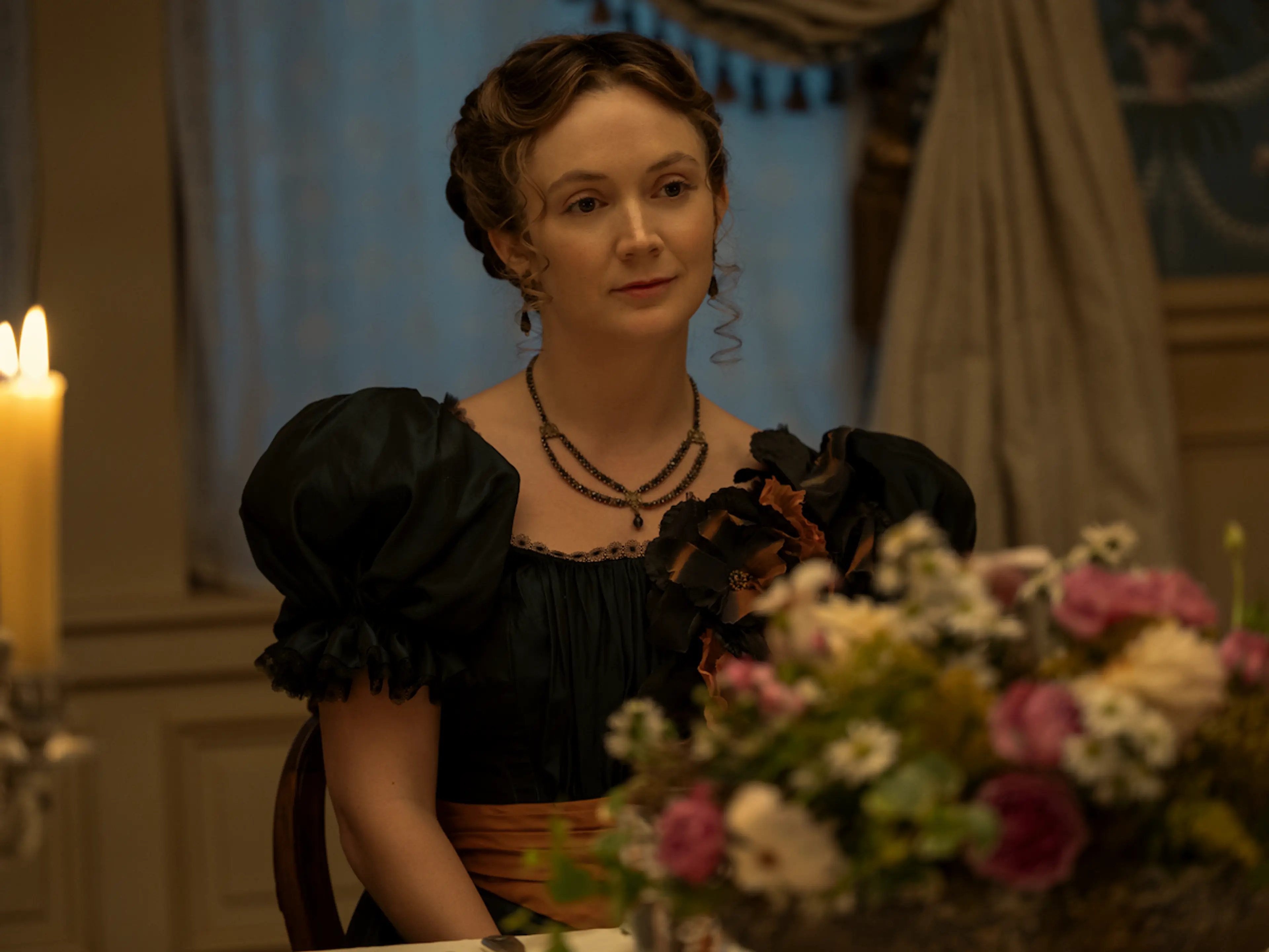 Billie Lourd as Emma Borden sits at a table with a flower arrangement in the foreground.