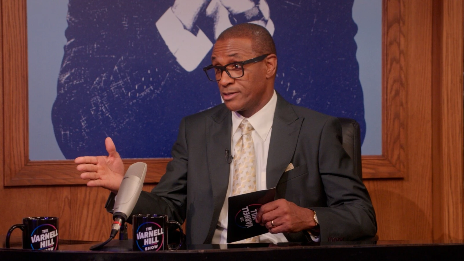Tommy Davidson appears as Varnell Hill behind a talk-show desk in an official Paramount+ still.