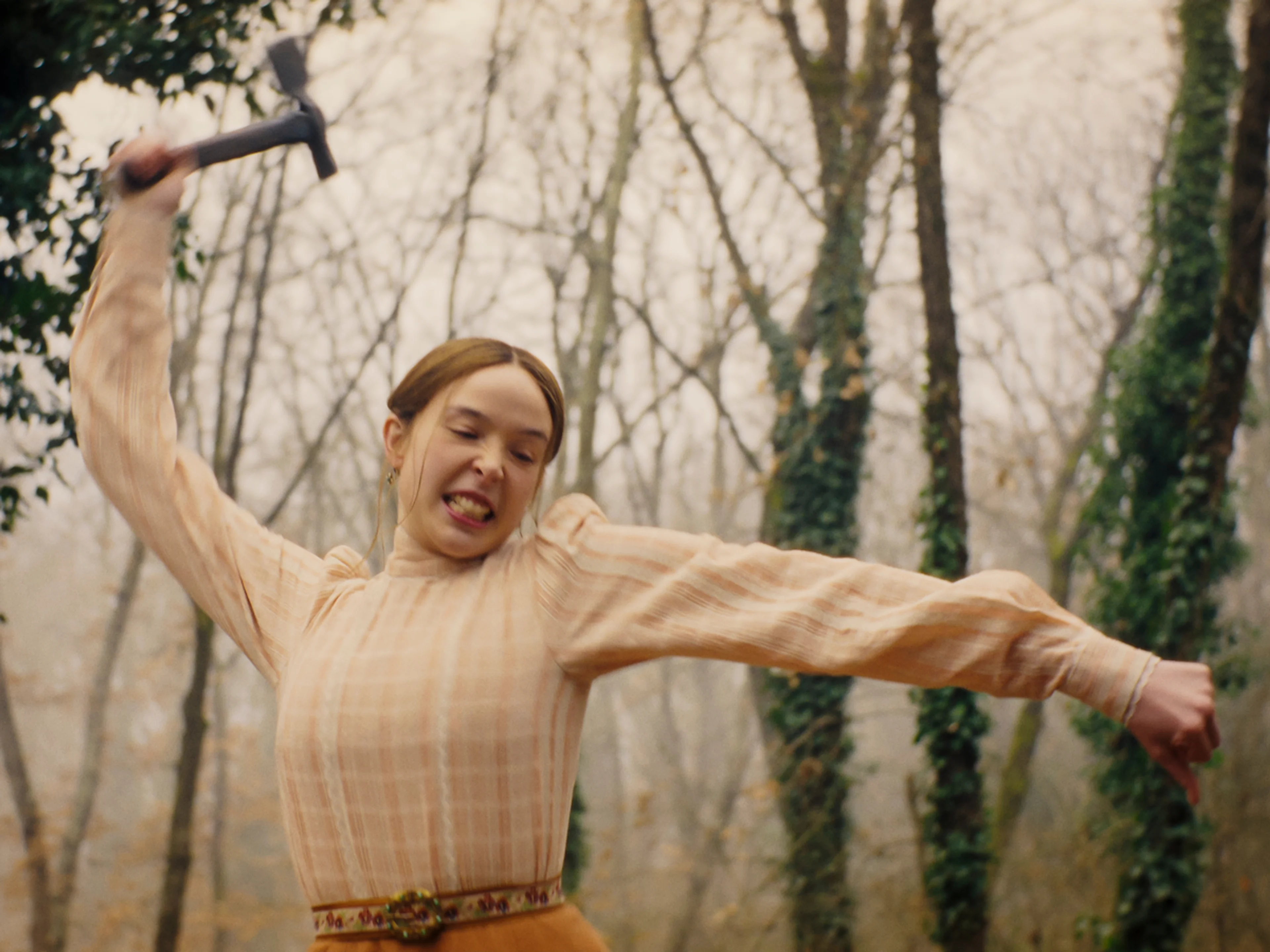 Ella Beatty as Lizzie Borden raises a hammer outdoors among the trees.