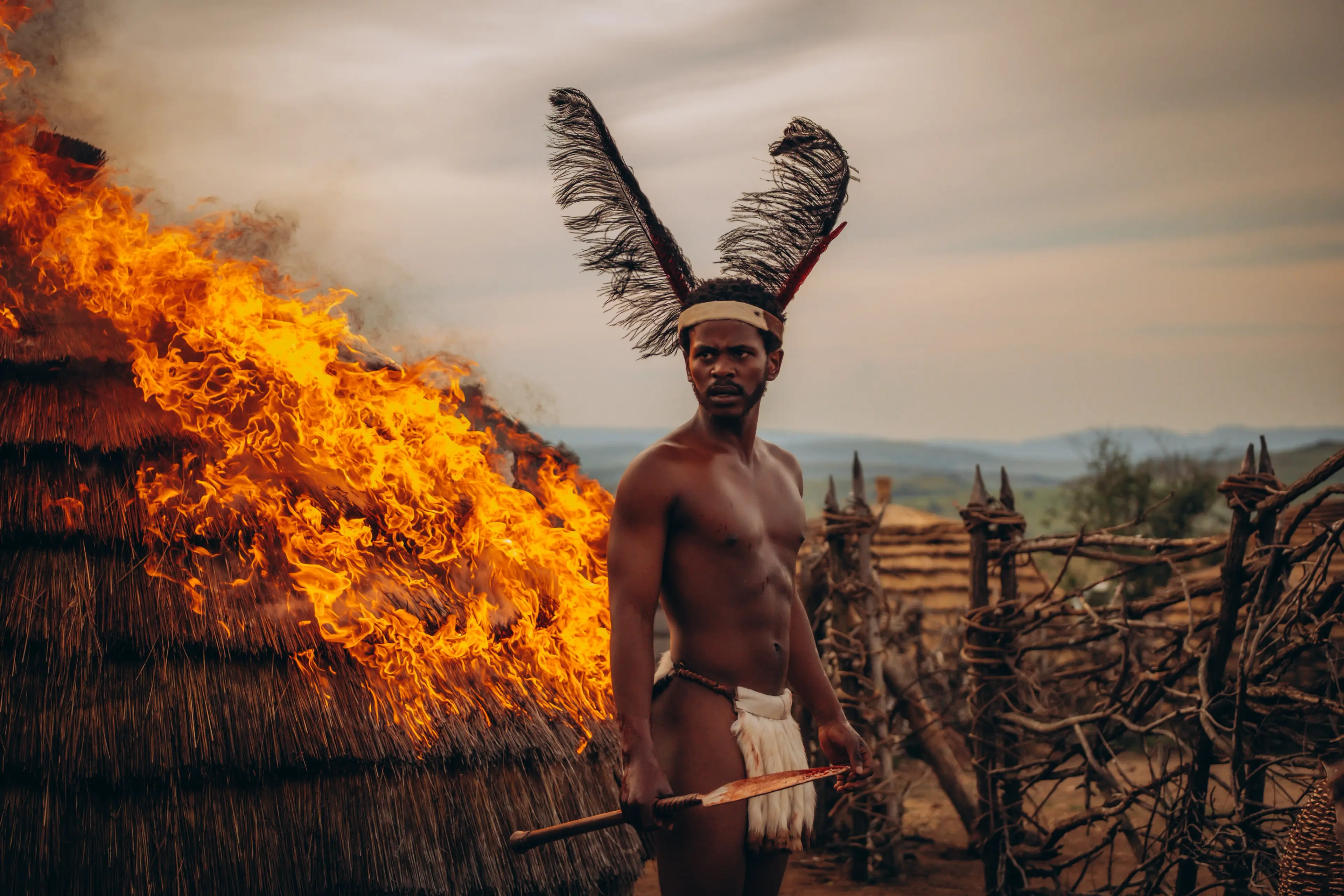 A man stands beside a burning hut in a Shaka iLembe Season 3 publicity image.