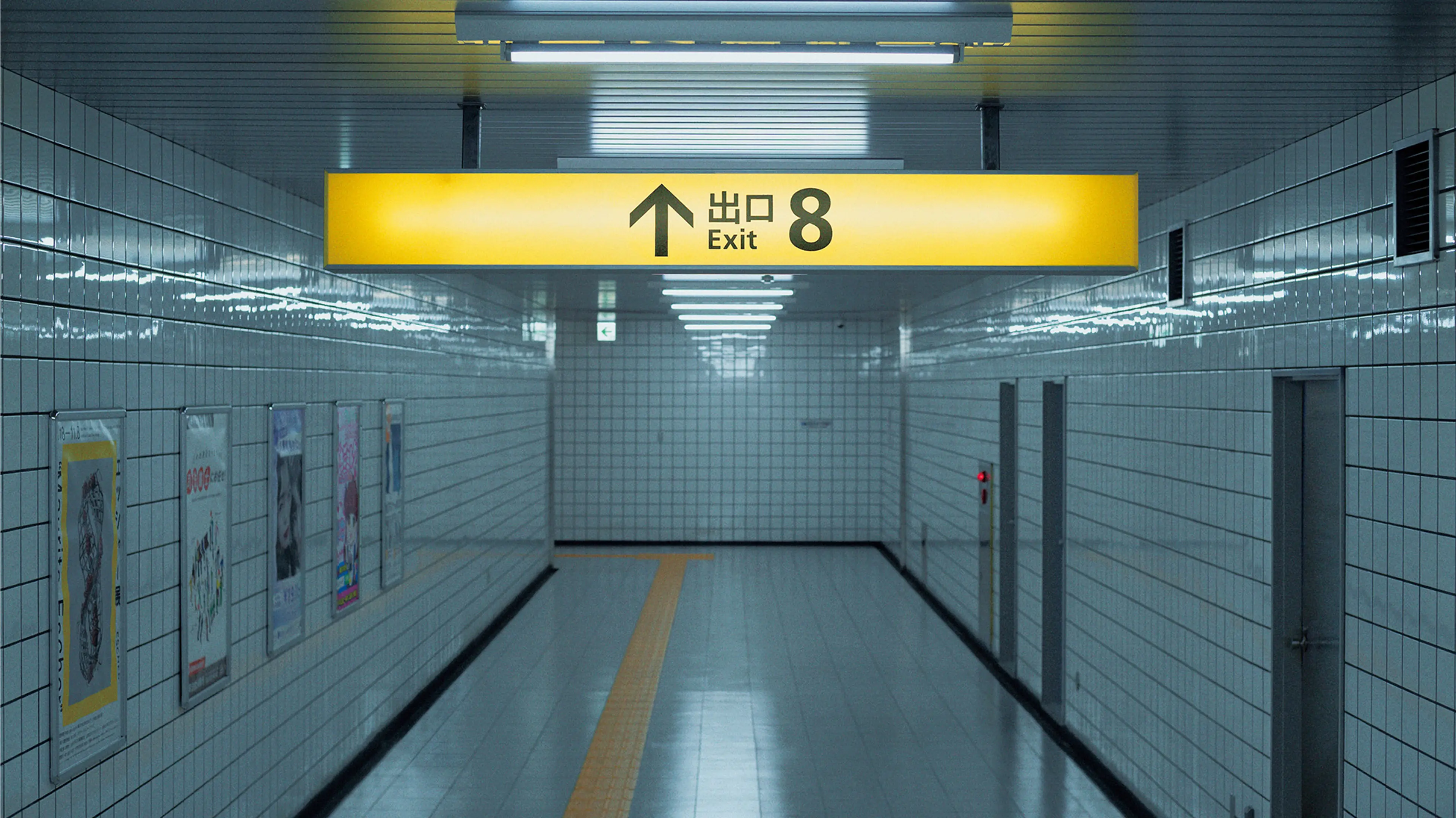 An empty white subway corridor has posters on the left wall and a yellow Exit 8 sign overhead.