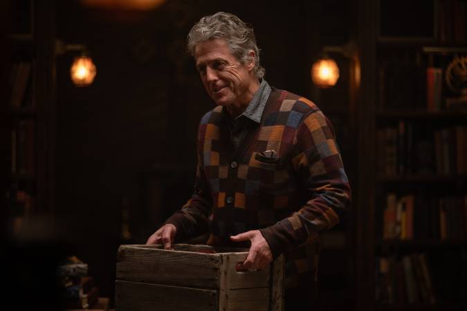 Hugh Grant as Reed smiles while standing indoors and holding a wooden crate.