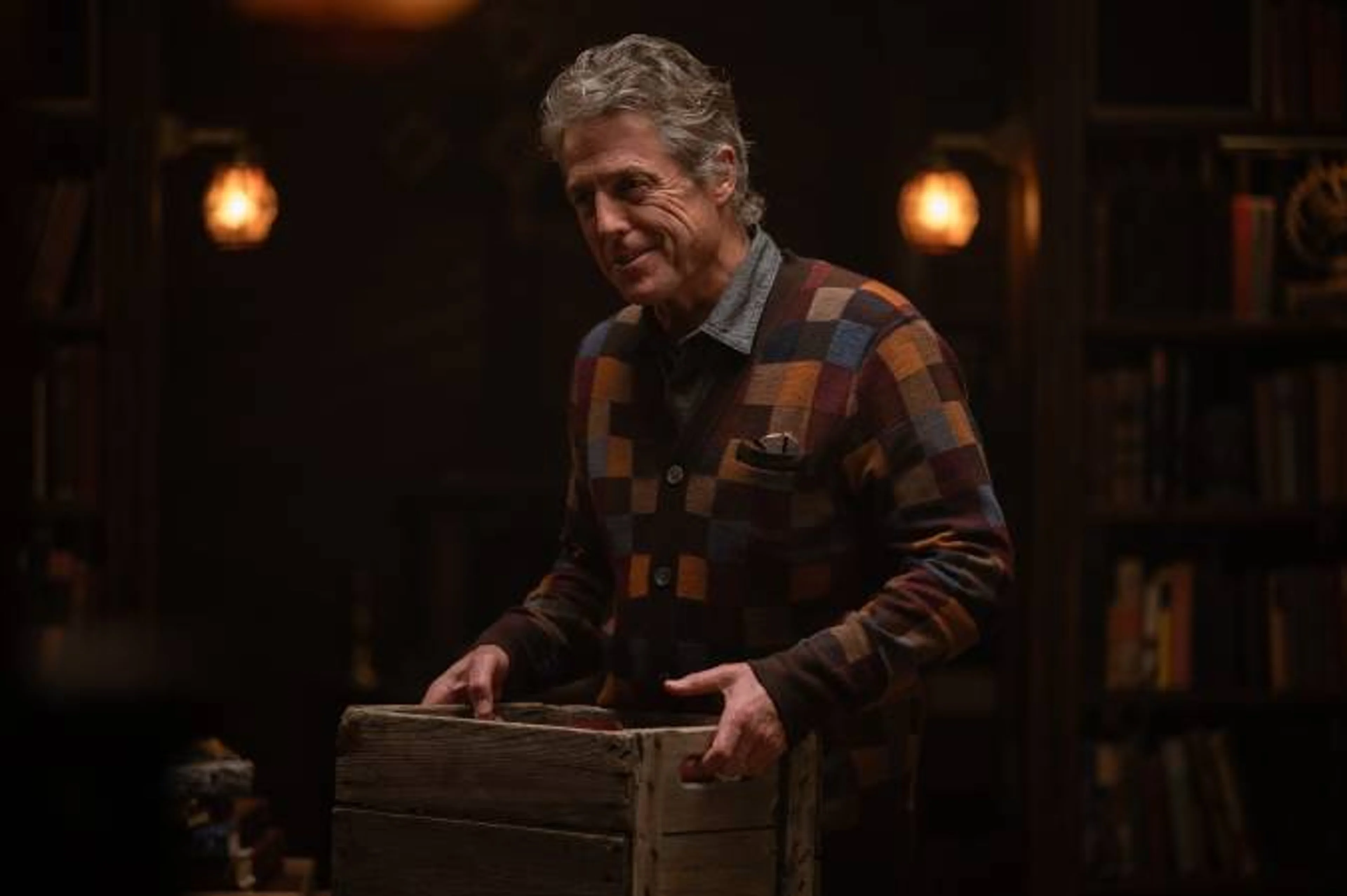 Hugh Grant as Reed smiles while standing indoors and holding a wooden crate.