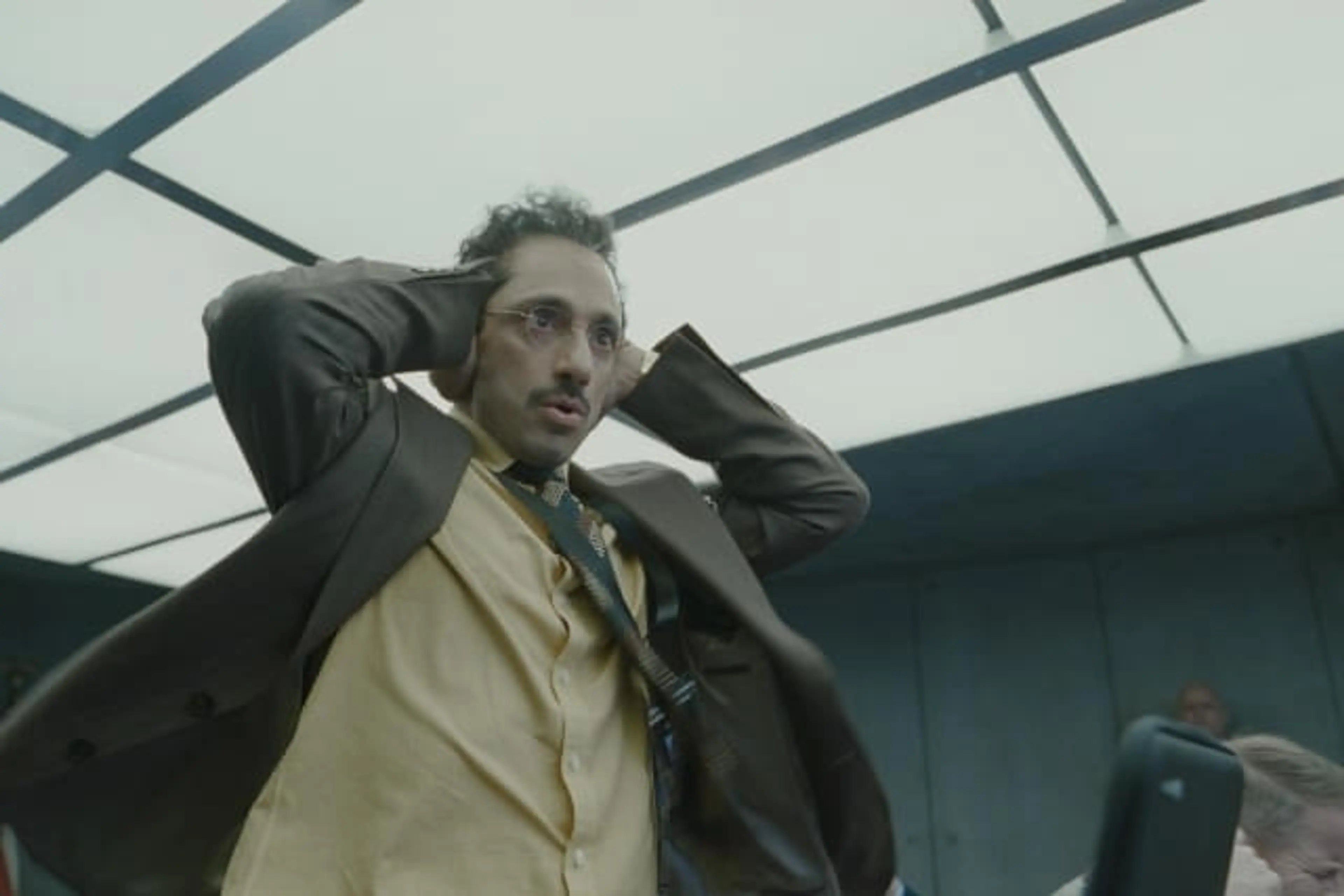 Riz Ahmed raises his hands beside his head beneath a glass ceiling in Digger.