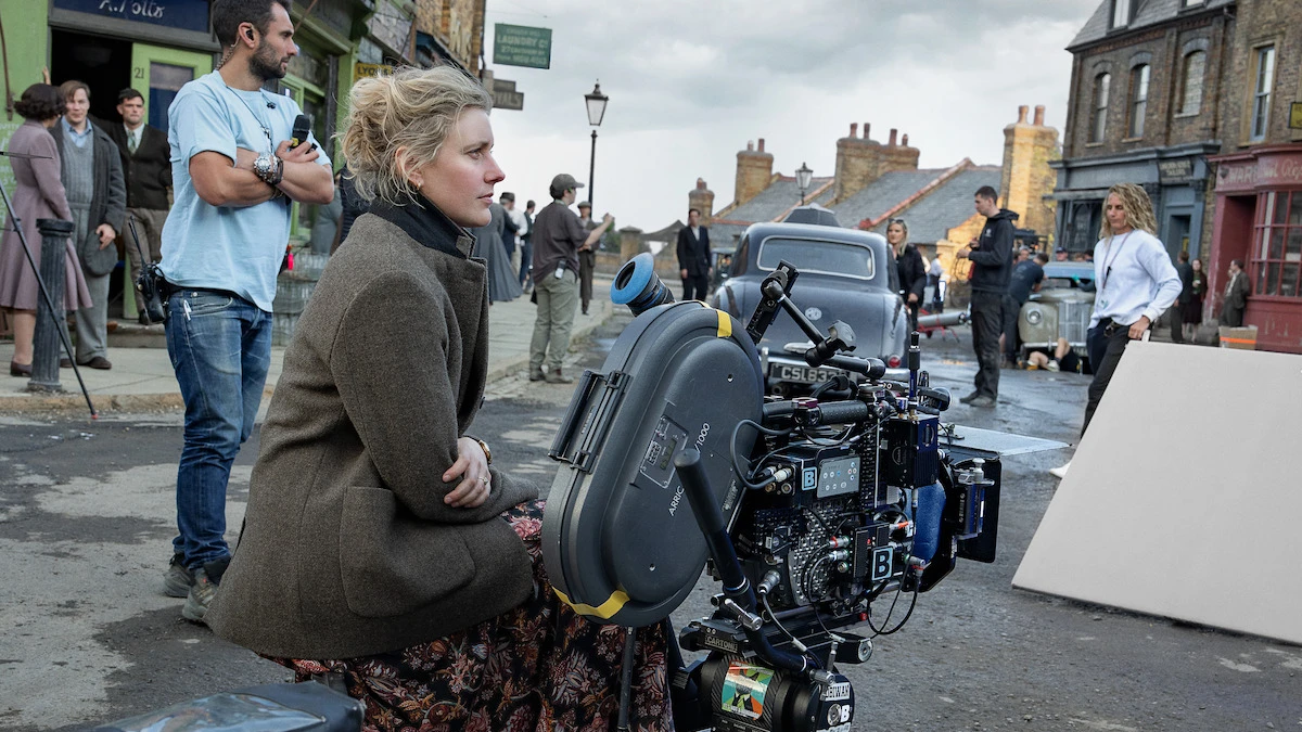 Greta Gerwig sits beside a large camera on the period London set of Narnia: The Magician’s Nephew.