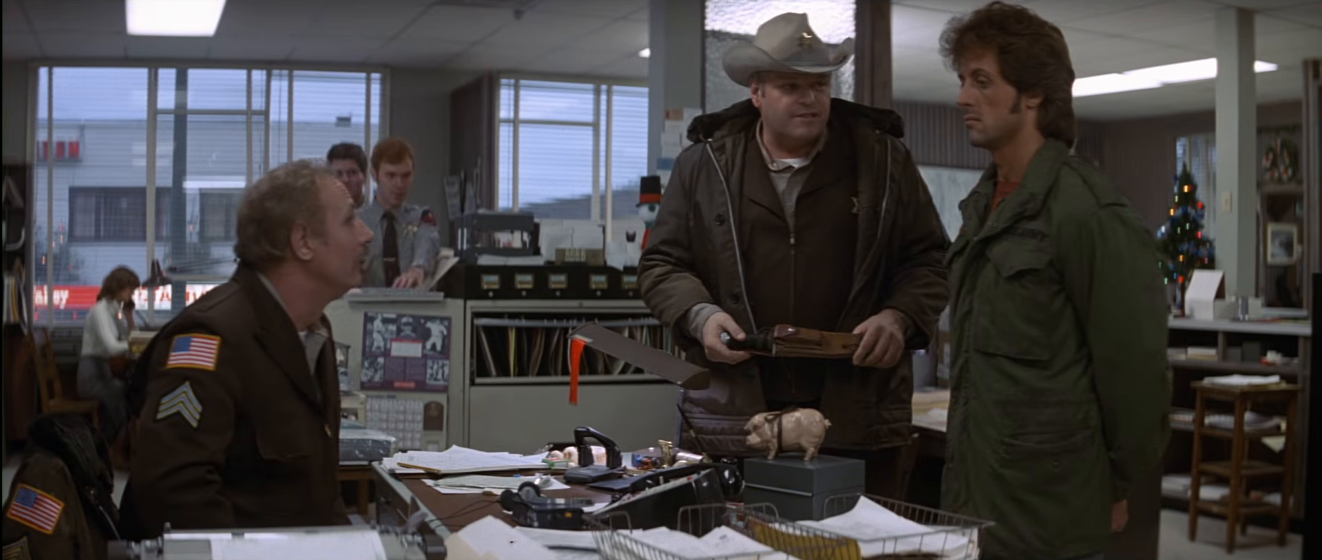 Rambo stands beside Sheriff Teasle, wearing a brown uniform and hat, in the police station.