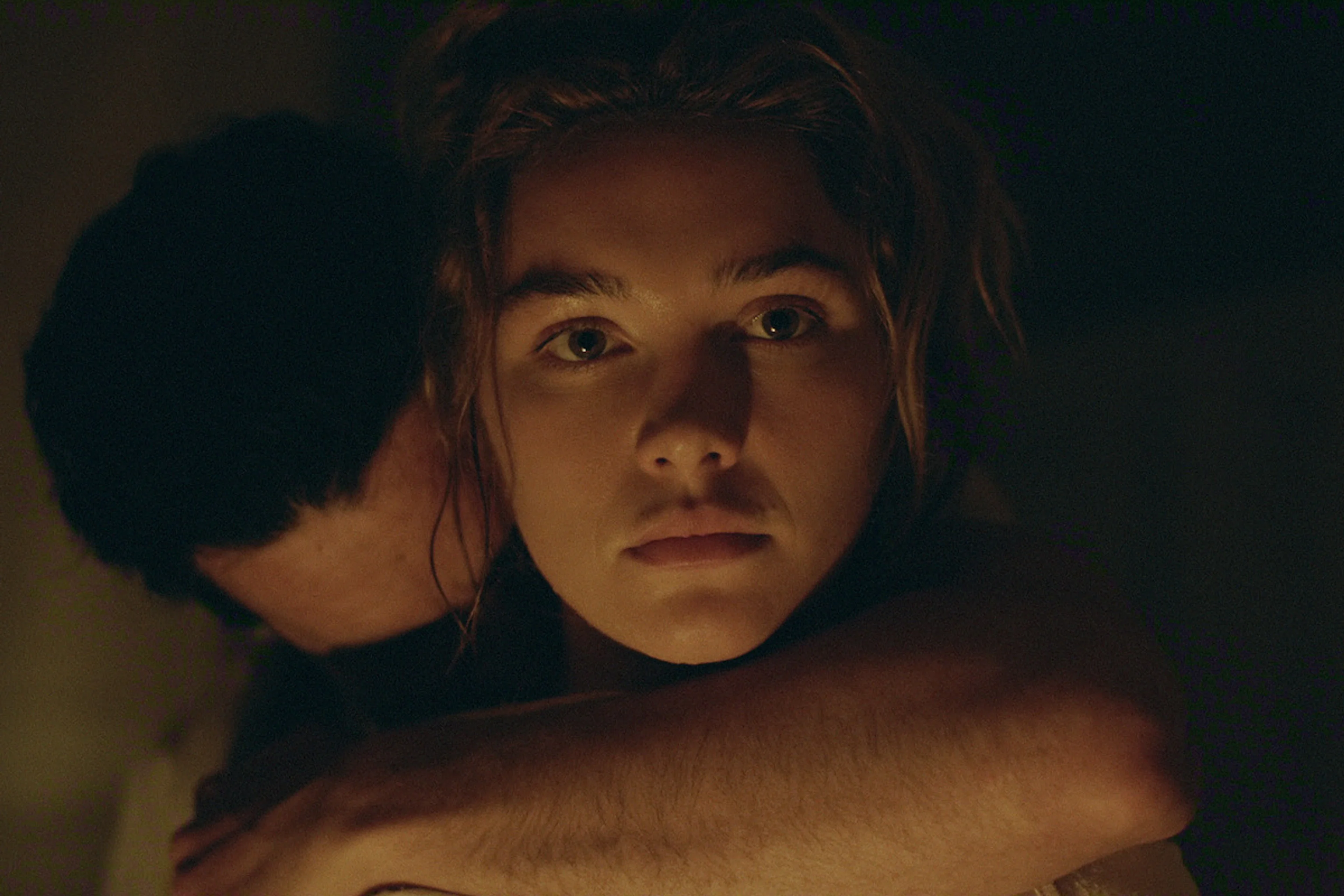 Cathy looks ahead while Adam embraces her from behind in a dark interior.