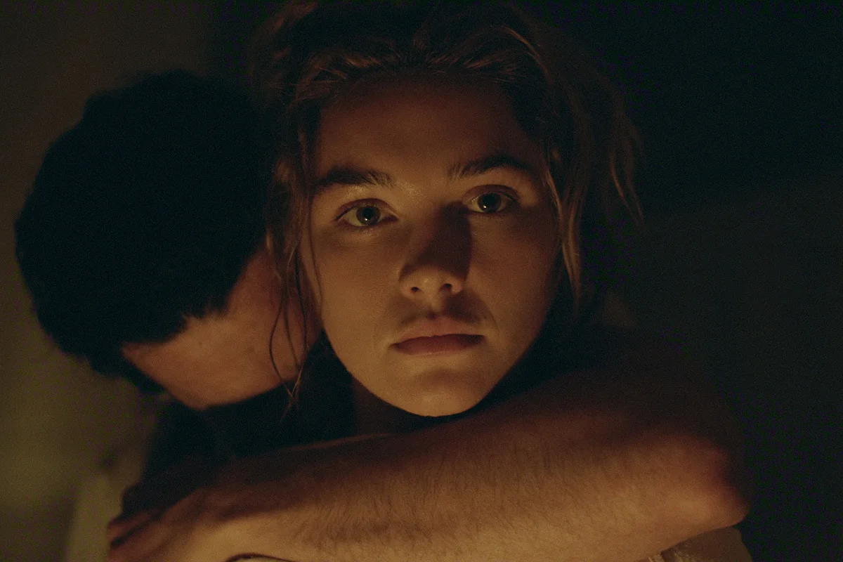 Cathy looks ahead while Adam embraces her from behind in a dark interior.