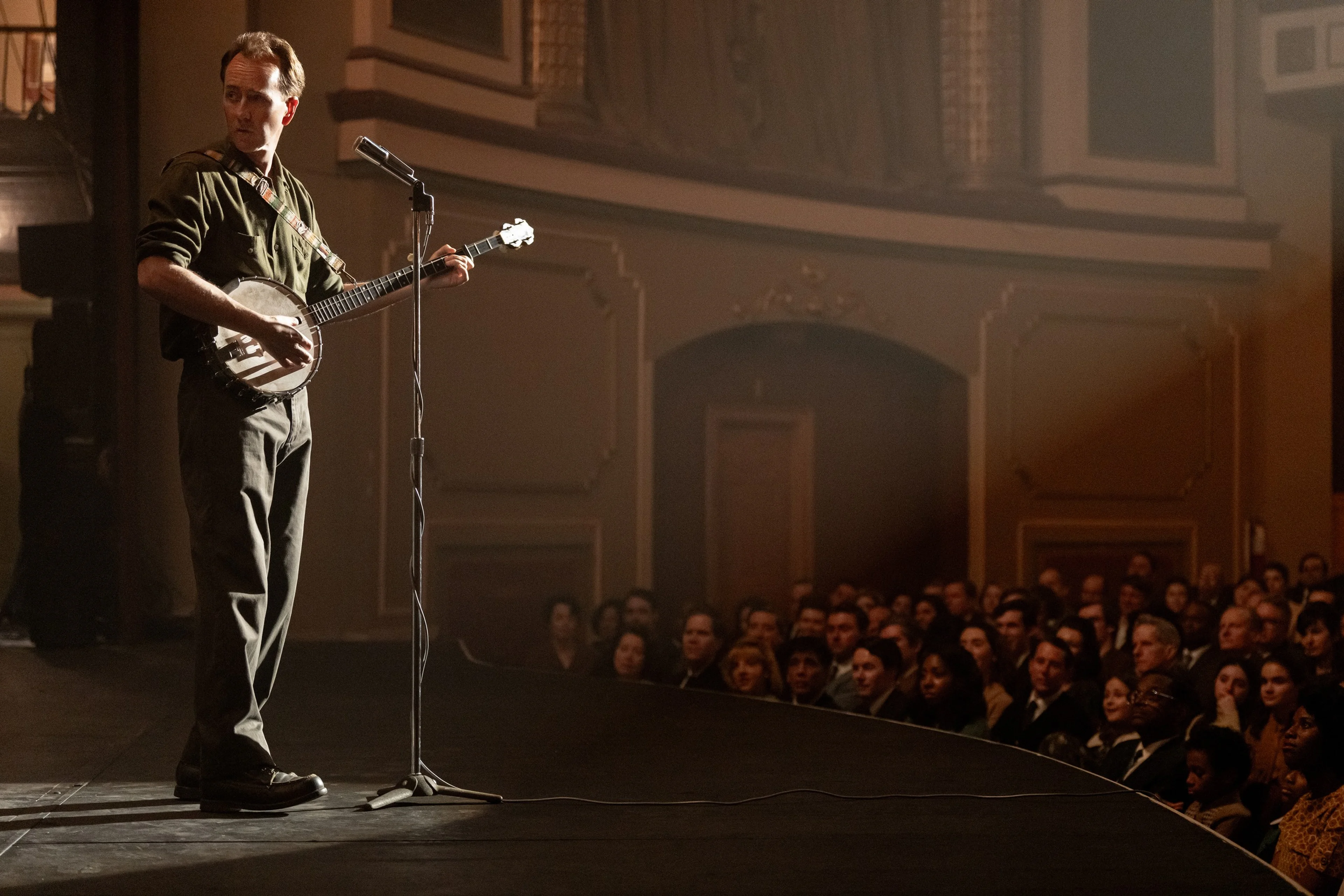 Edward Norton as Pete Seeger holds a banjo on a stage, with audience members visible beside him.