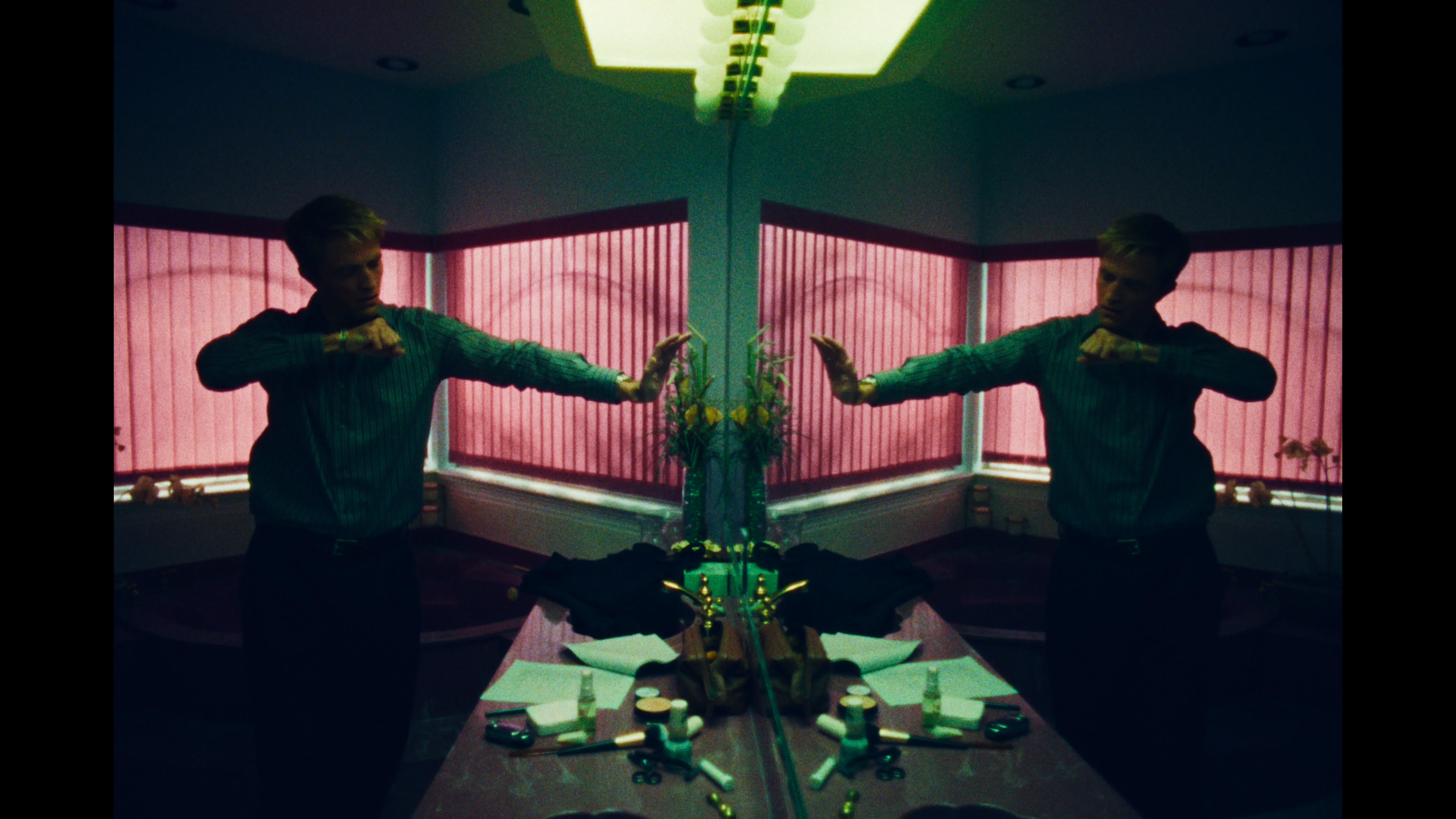 Pattinson stands before a mirror with his hands raised under green light and pink blinds.