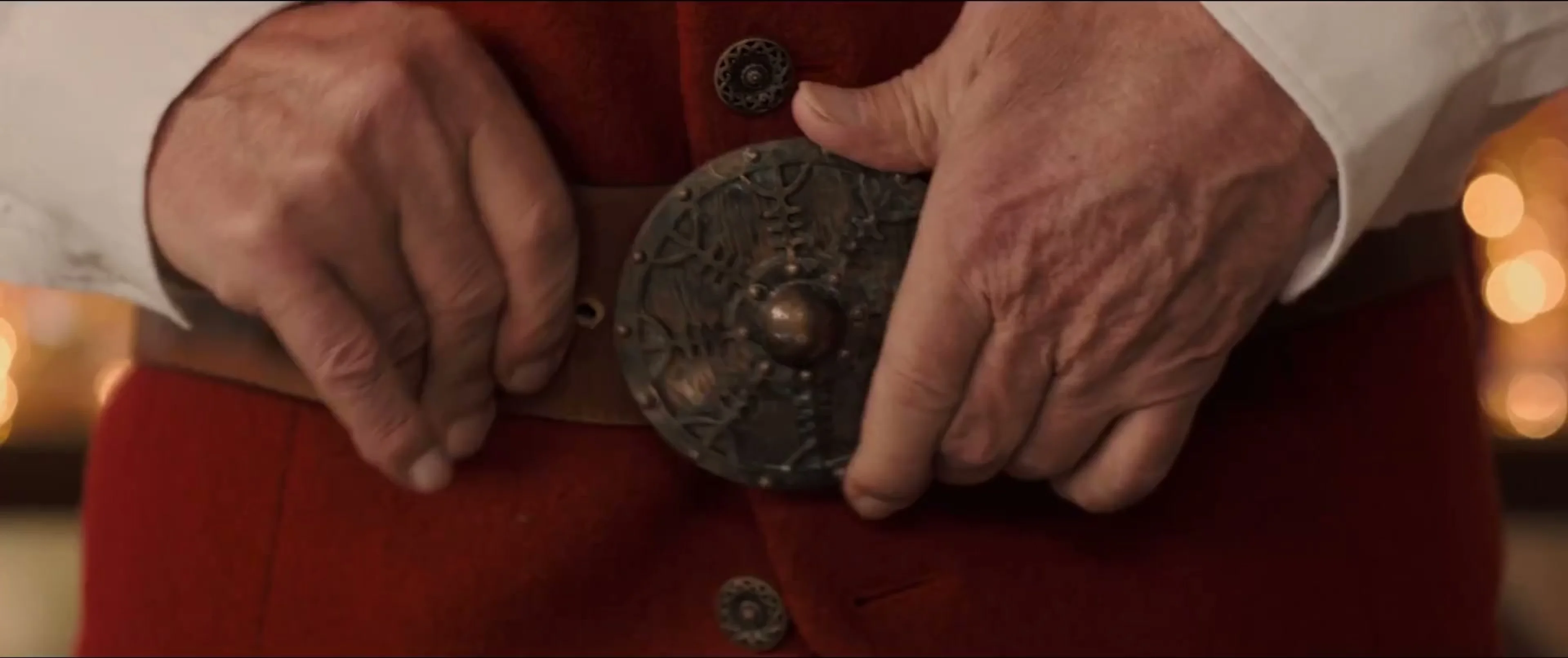 Santa’s bare hands rest against his red suit beside an ornate metal belt.