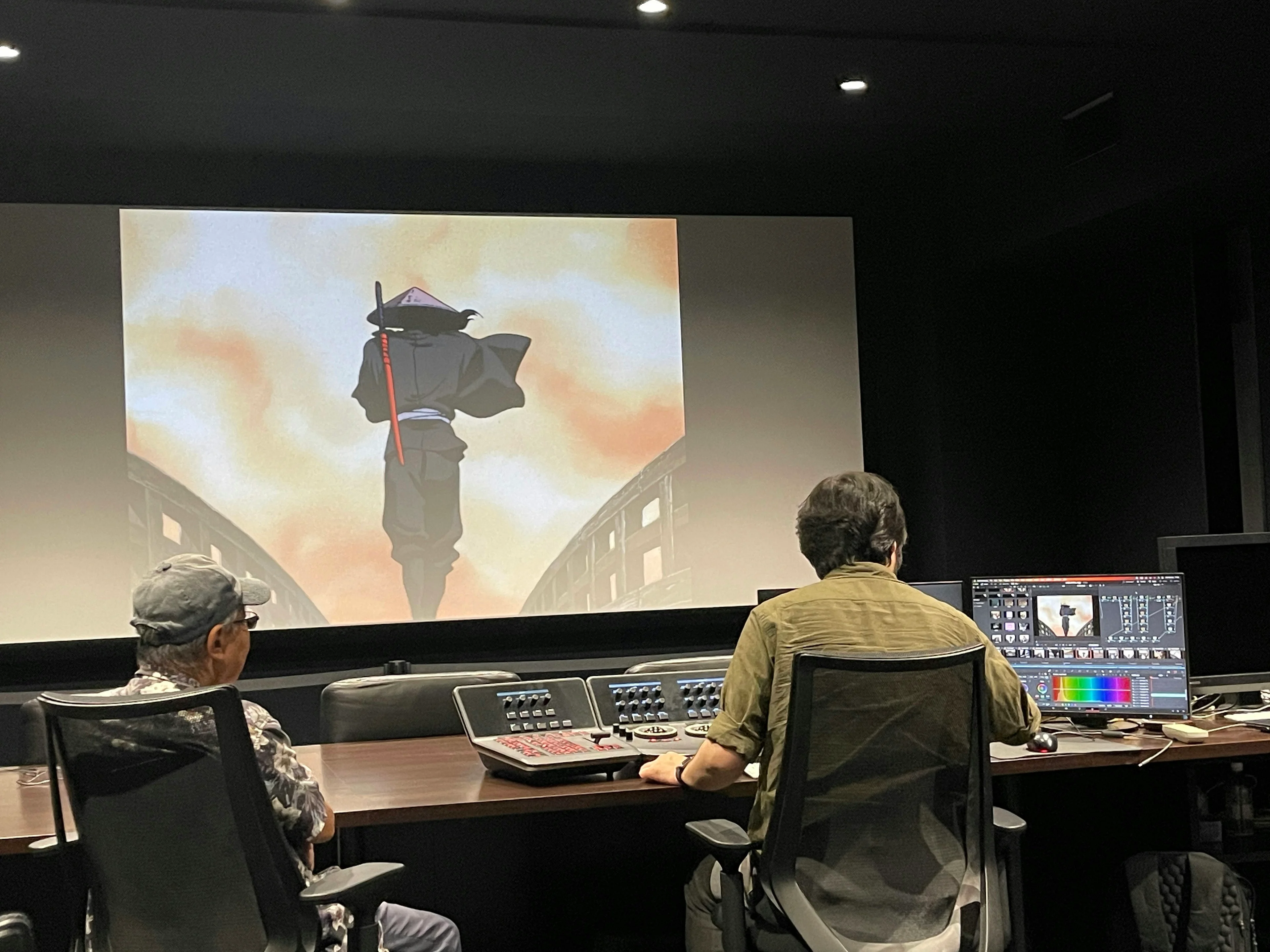 Two people watch Jubei projected on a large screen beside a color-correction monitor and control desk.