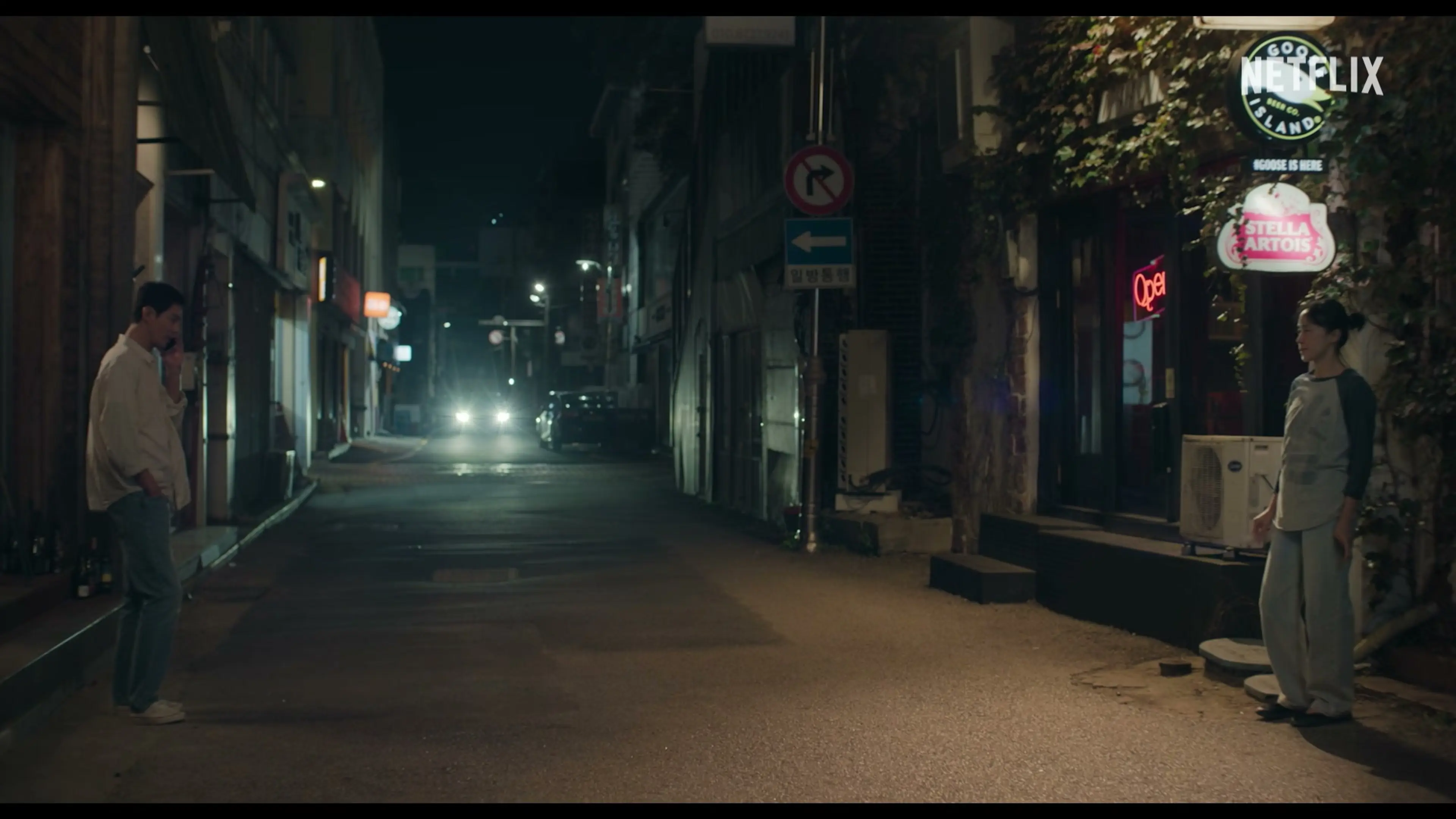 Sang-woo uses his phone on one side of a street while Mi-ok stands on the other.