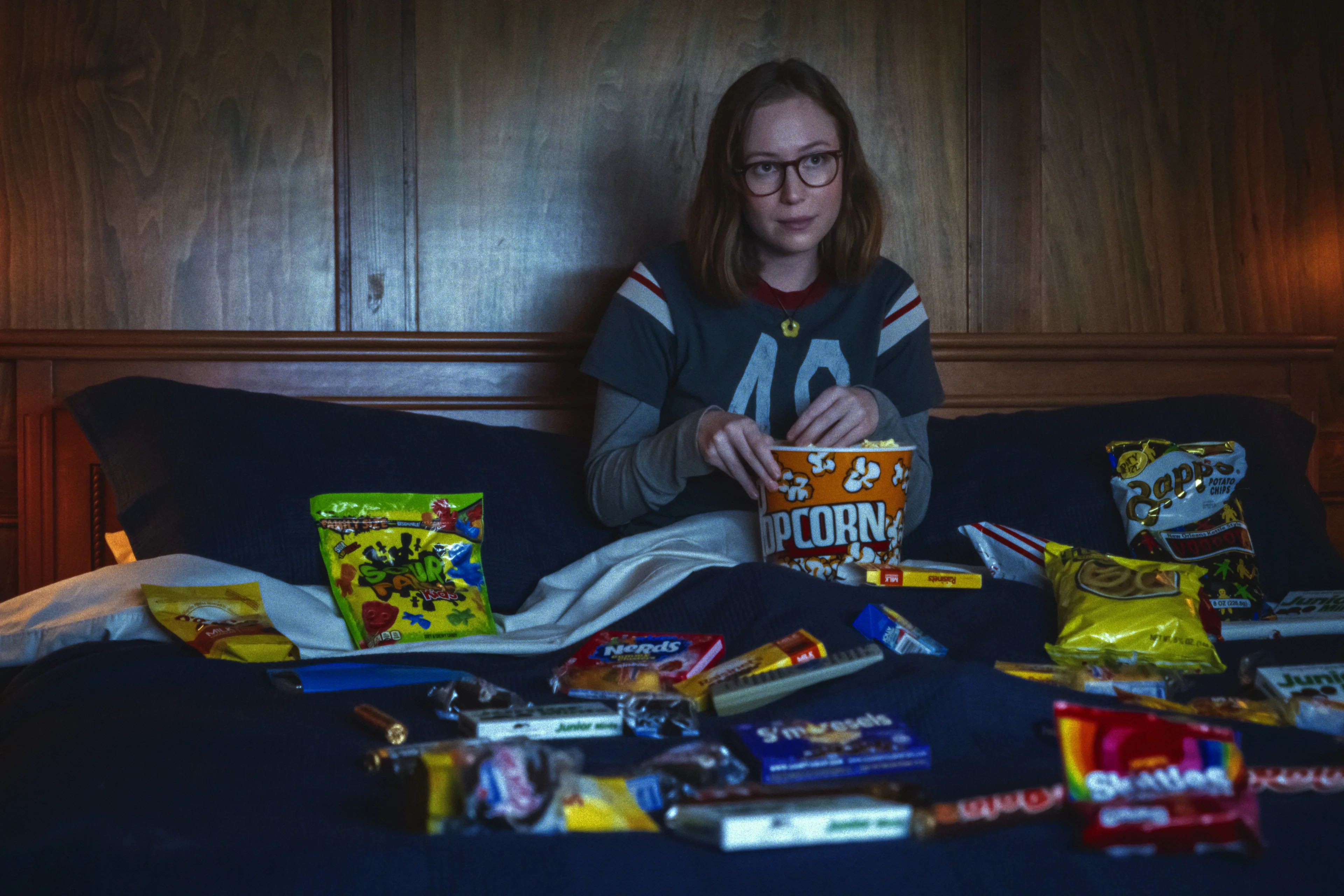 Hannah Einbinder as Kris sits with popcorn and several bags of snacks.