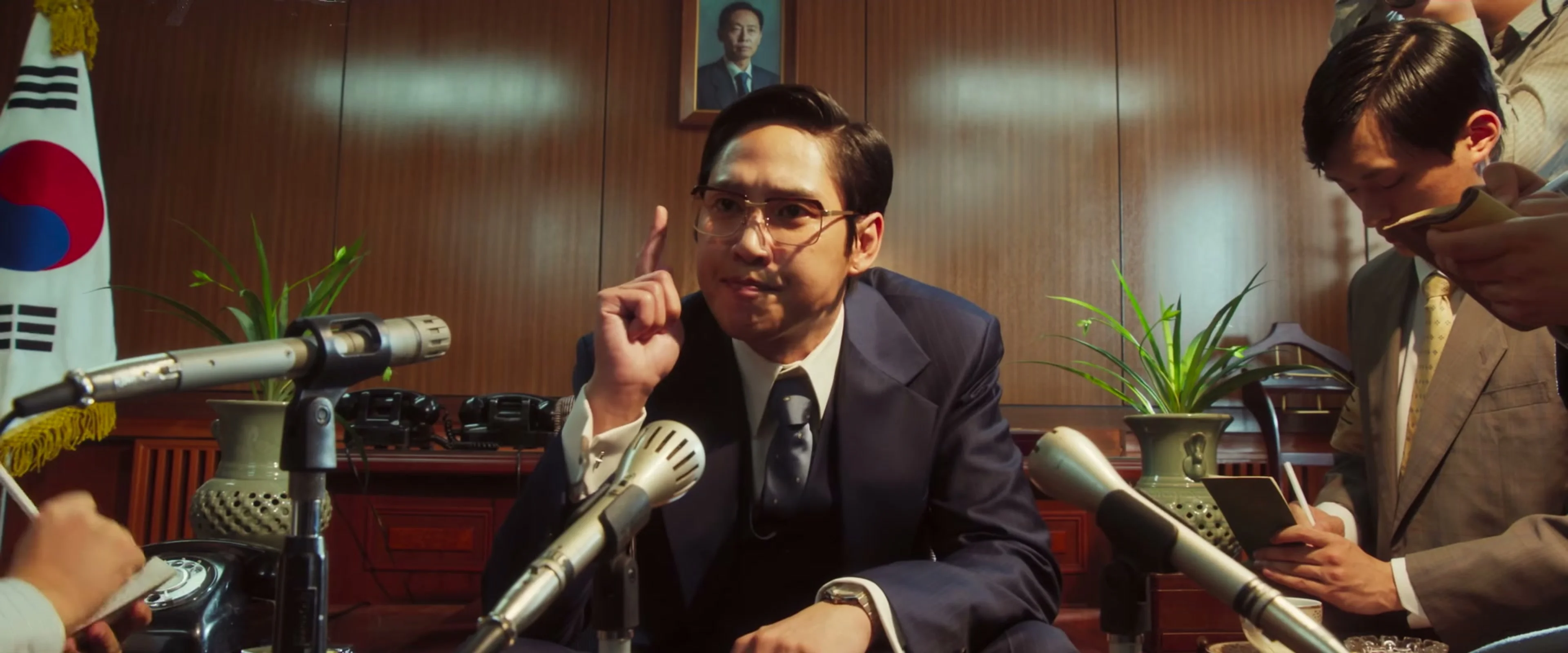 A suited man addresses microphones in a sharp frame from The Assassin(s) trailer.