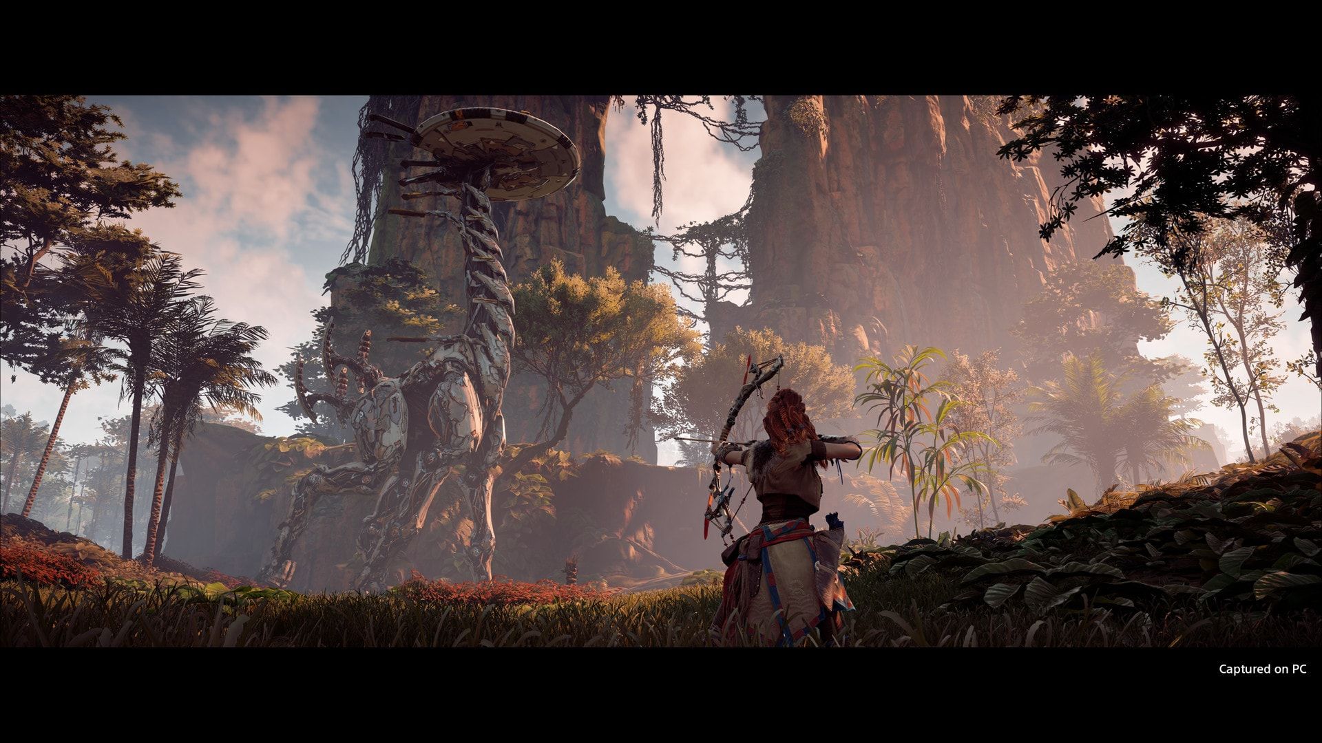 Aloy stands below a giant machine in a wooded landscape.