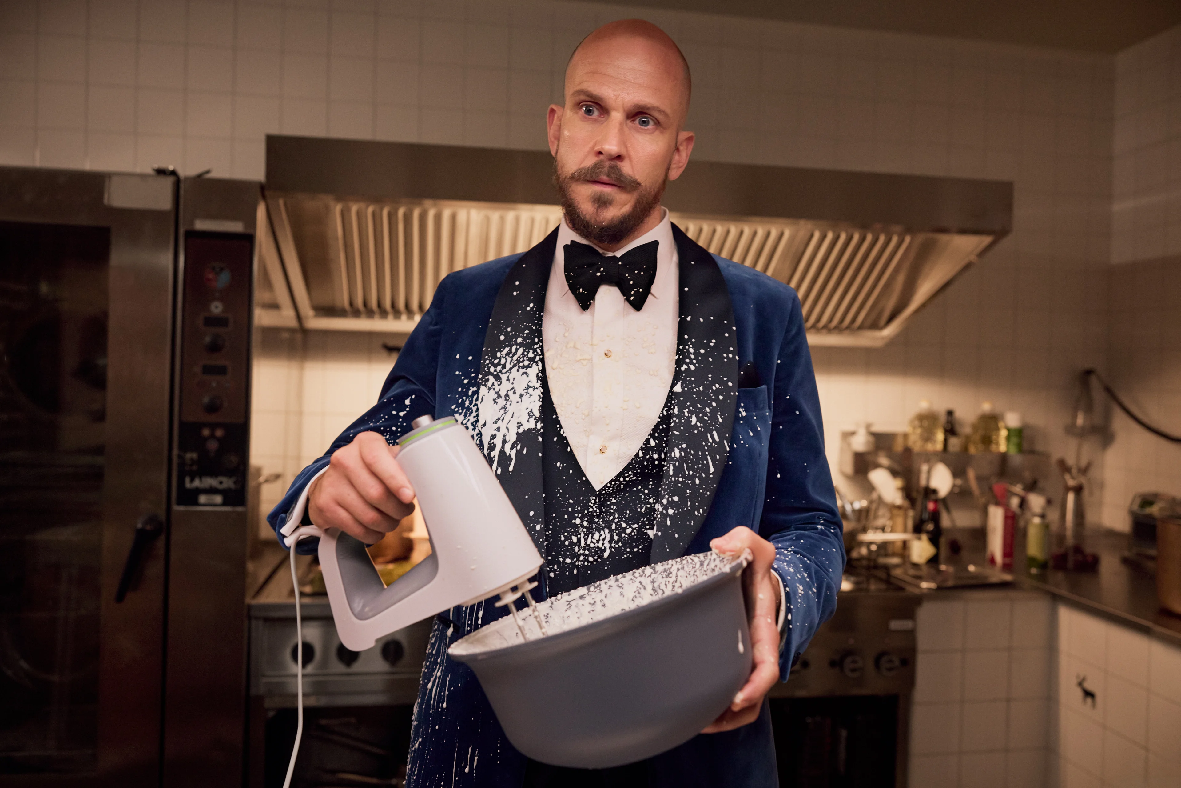 A man in evening clothes holds a mixing bowl in a flour-spattered kitchen.