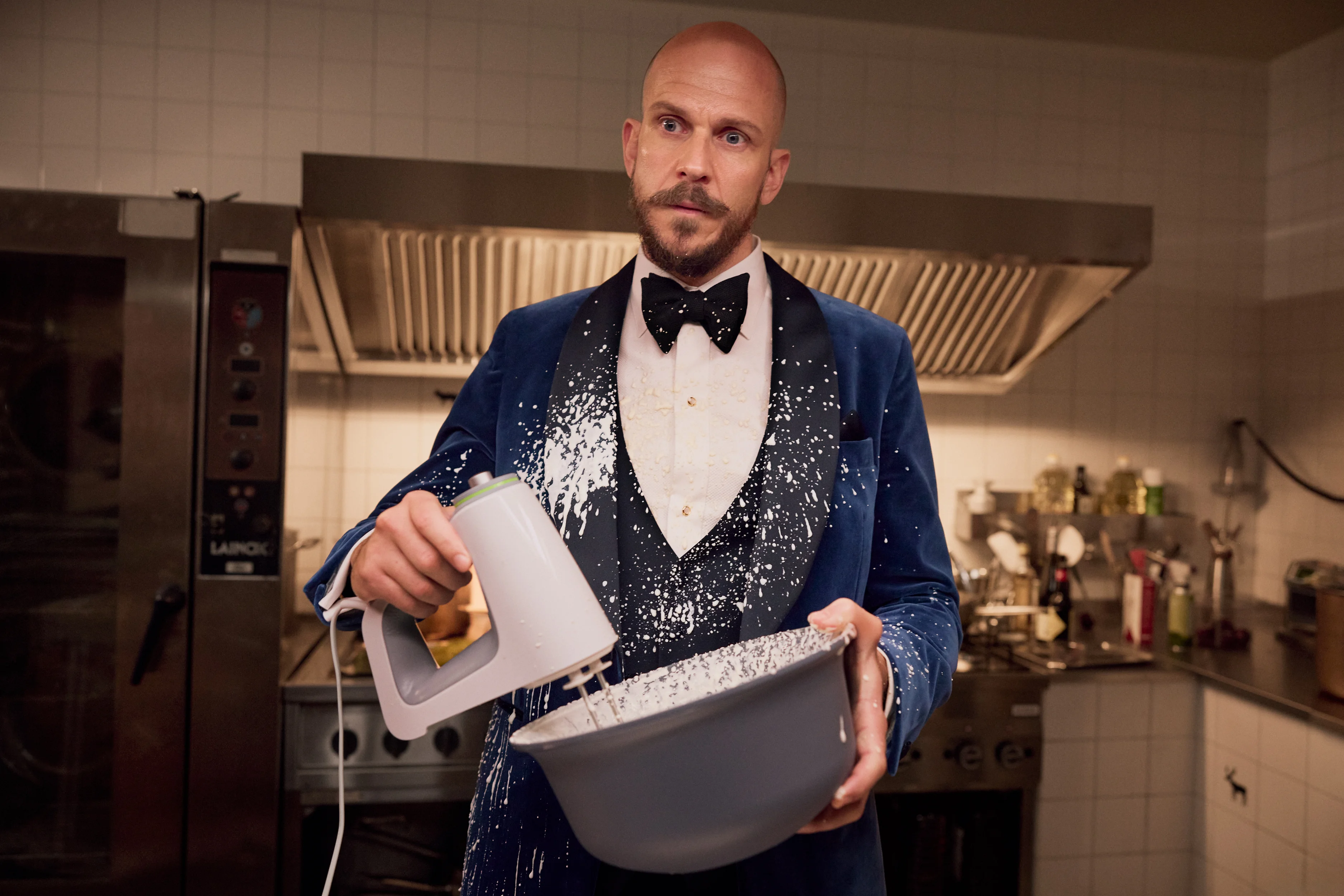 A man in evening clothes holds a mixing bowl in a flour-spattered kitchen.