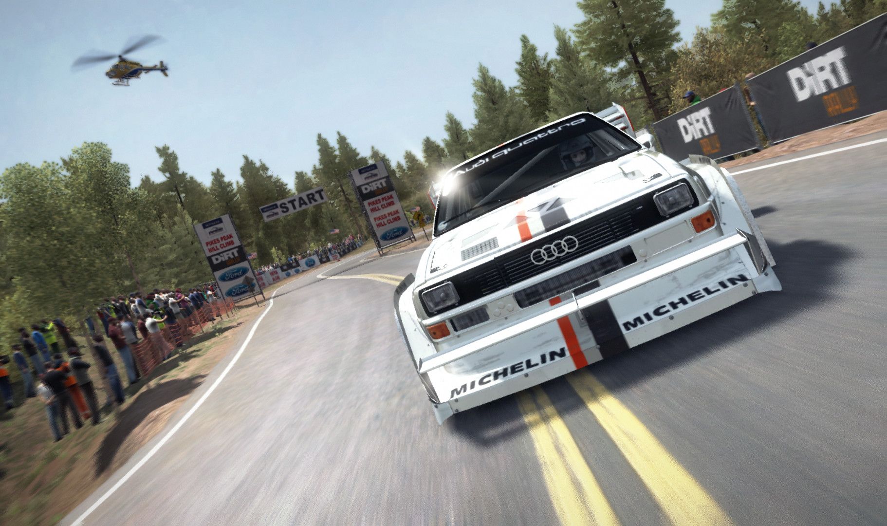 White Audi rally car drives around a paved corner with spectators and a helicopter nearby.