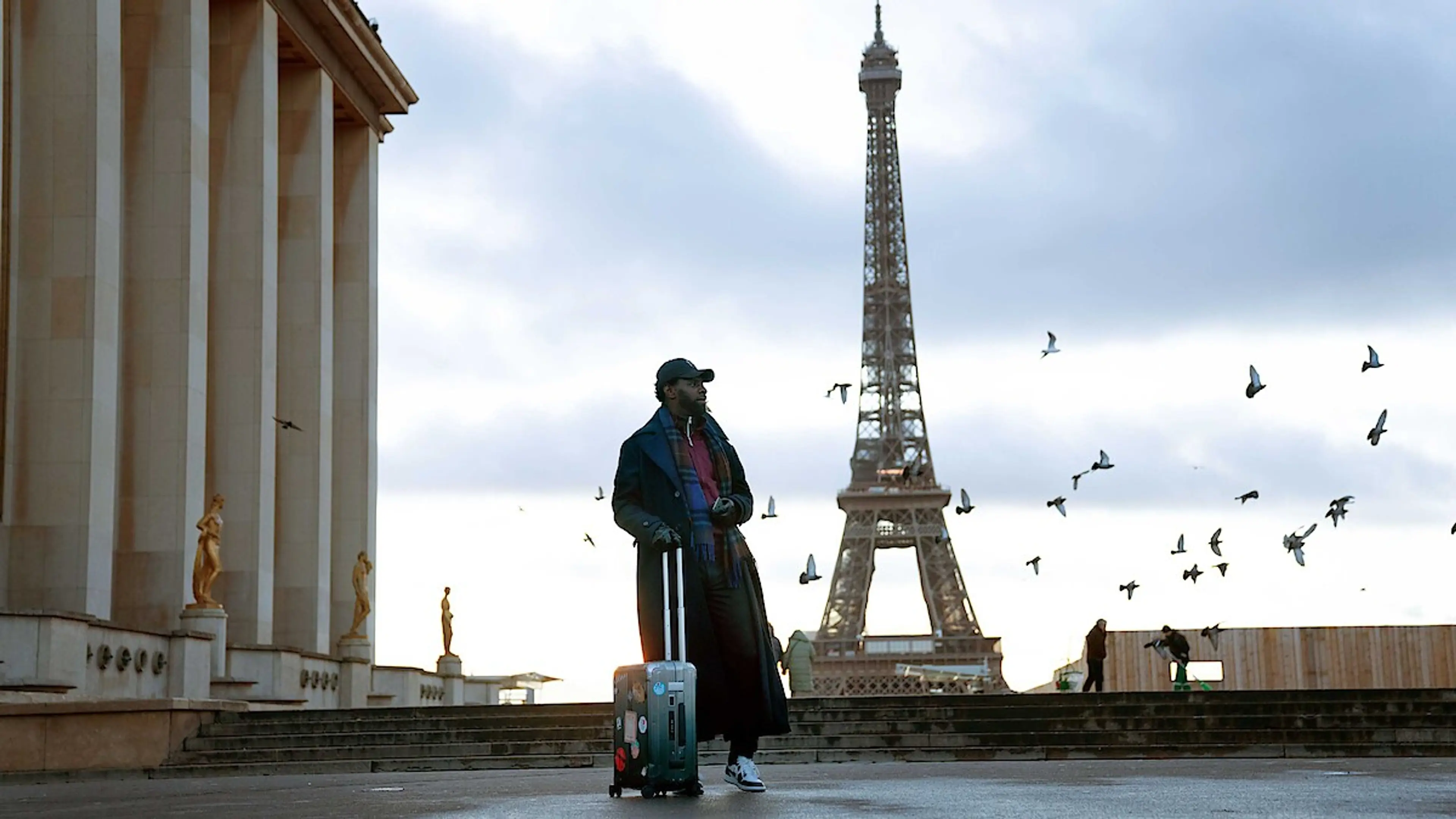Omar Sy as Assane Diop stands with a suitcase in front of the Eiffel Tower in Lupin.