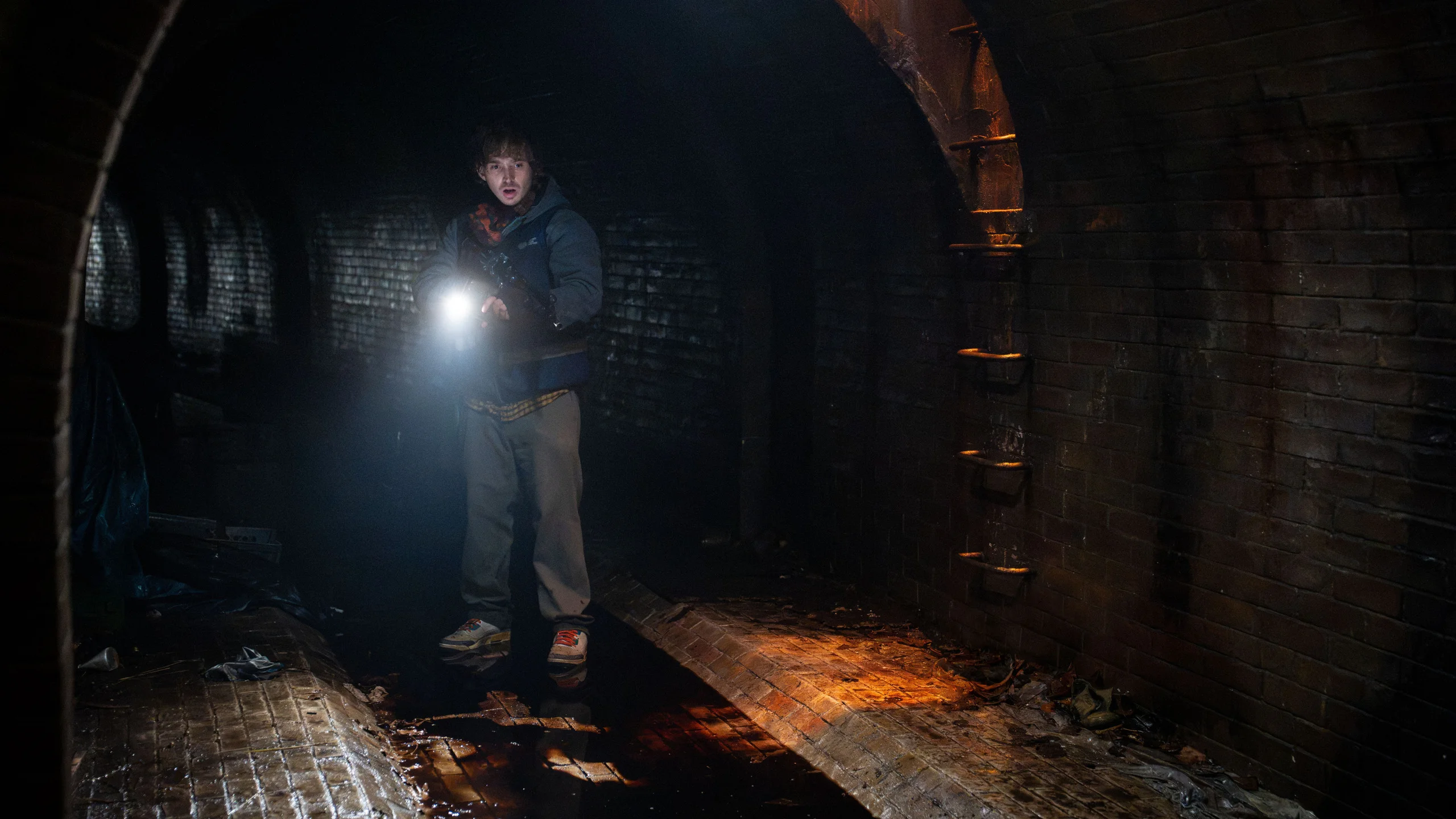 Austin Abrams as Bryan holds a flashlight inside a dark brick tunnel in Resident Evil.