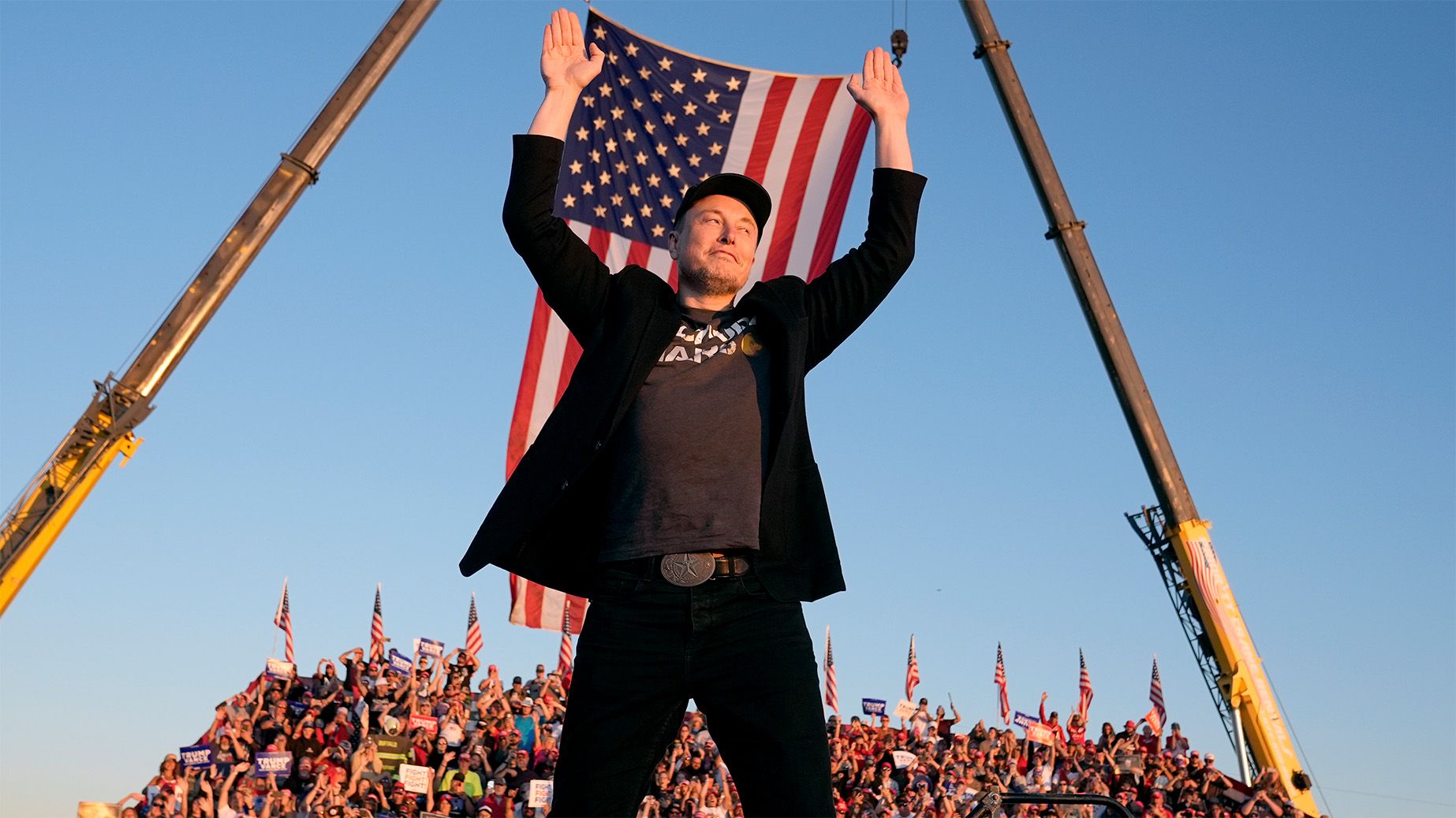 Elon Musk raises both hands in front of a large United States flag at an outdoor event.