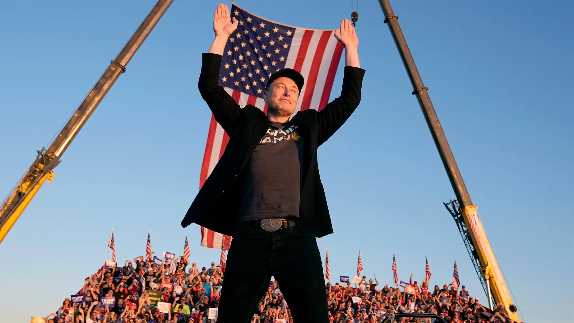 Elon Musk raises both hands in front of a large United States flag at an outdoor event.