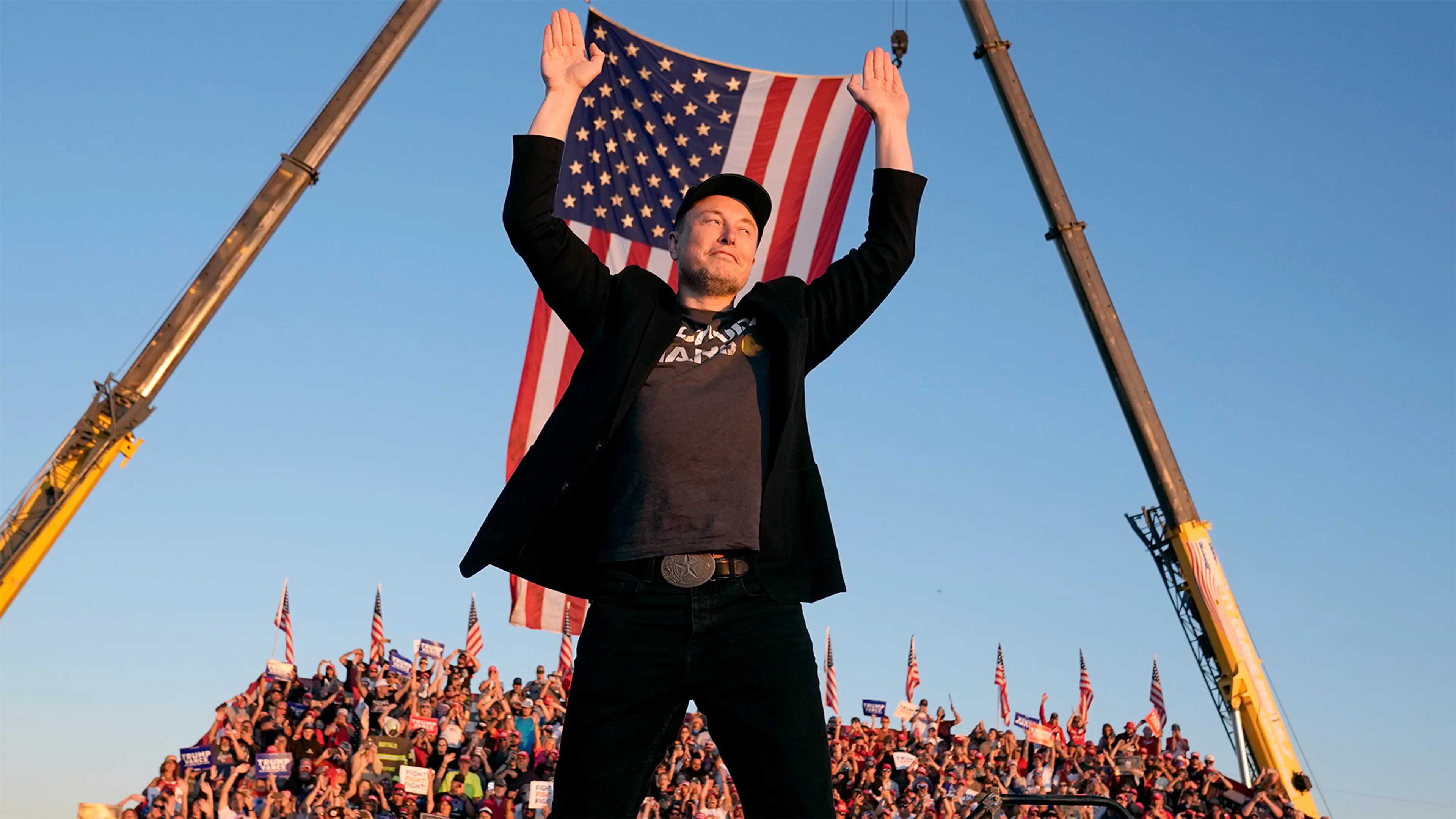 Elon Musk raises both hands in front of a large United States flag at an outdoor event.