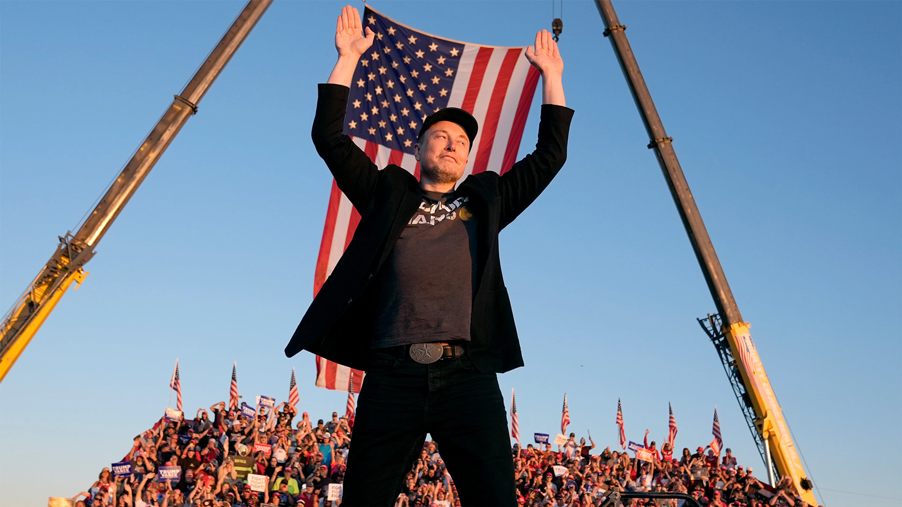 Elon Musk raises both hands in front of a large United States flag at an outdoor event.