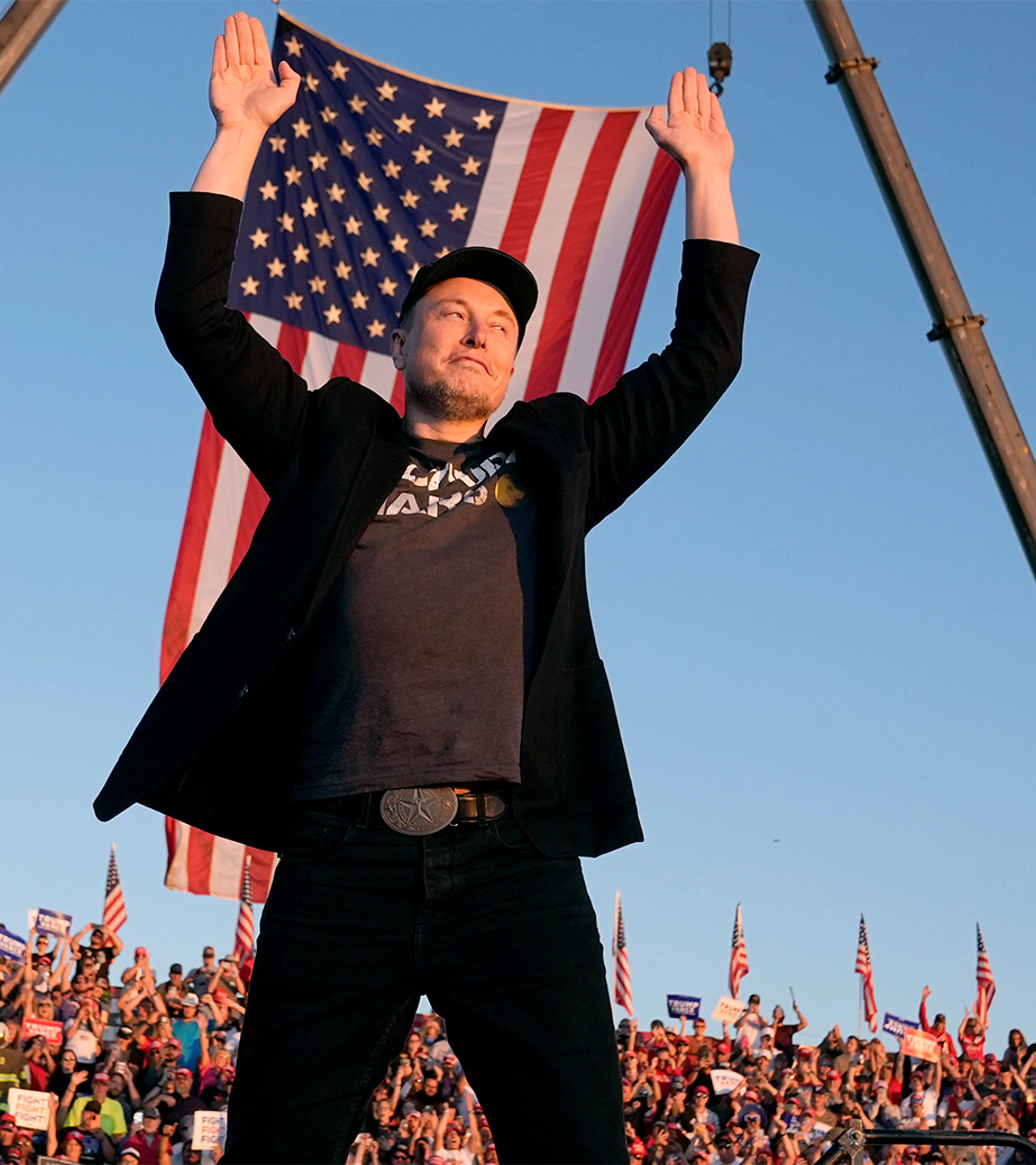 Elon Musk raises both hands in front of a large United States flag at an outdoor event.