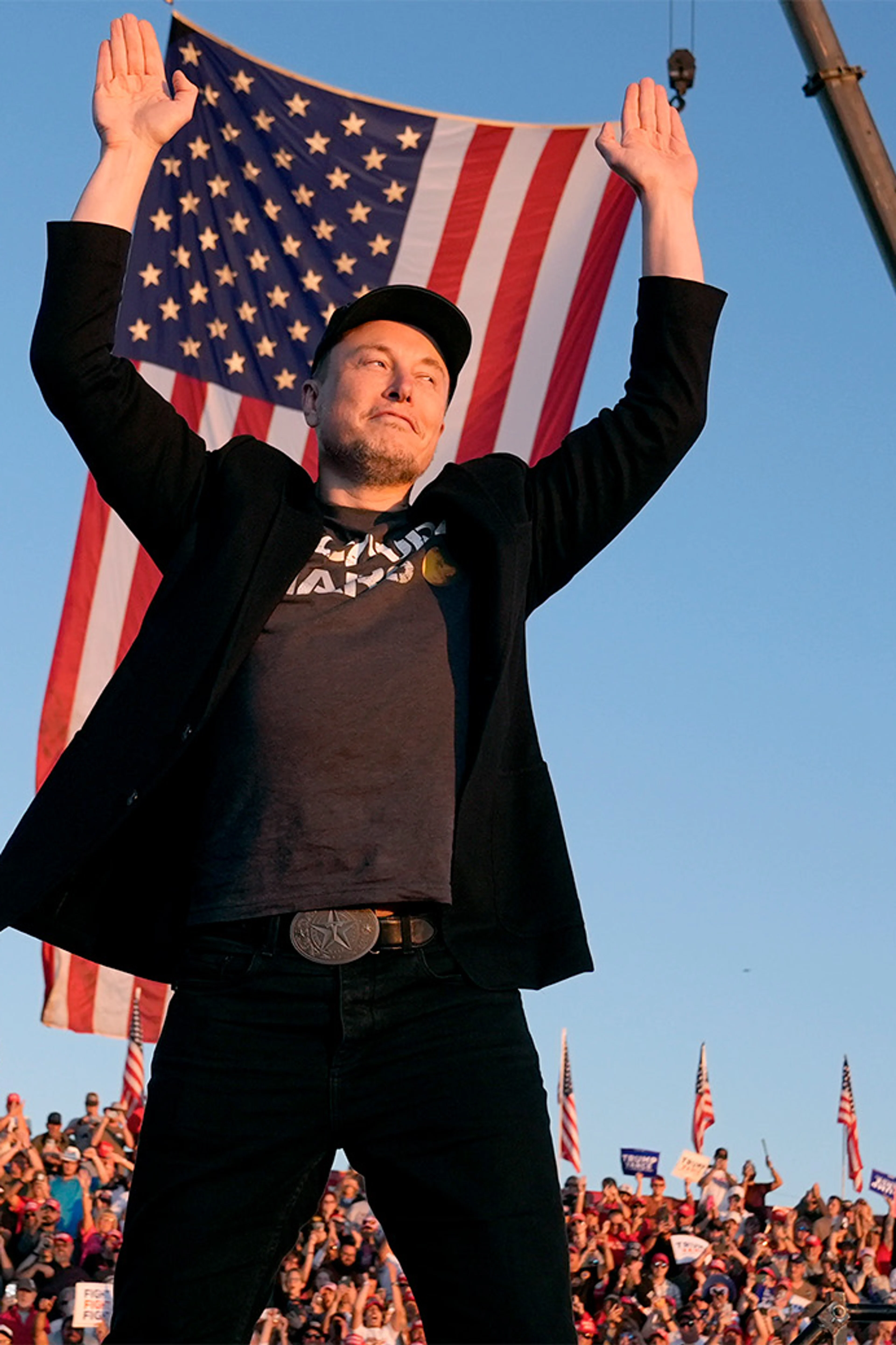 Elon Musk raises both hands in front of a large United States flag at an outdoor event.
