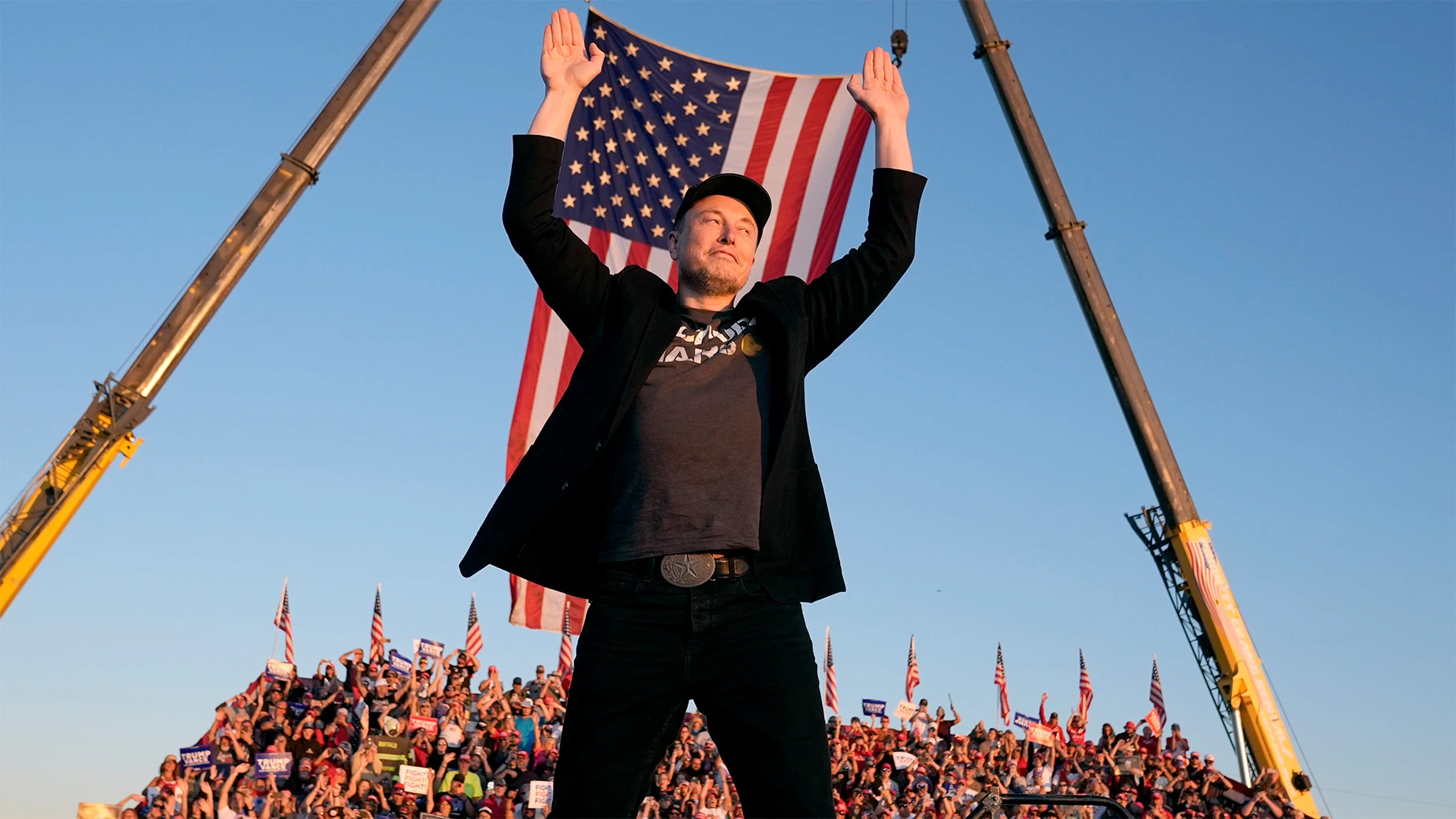 Elon Musk raises both hands in front of a large United States flag at an outdoor event.
