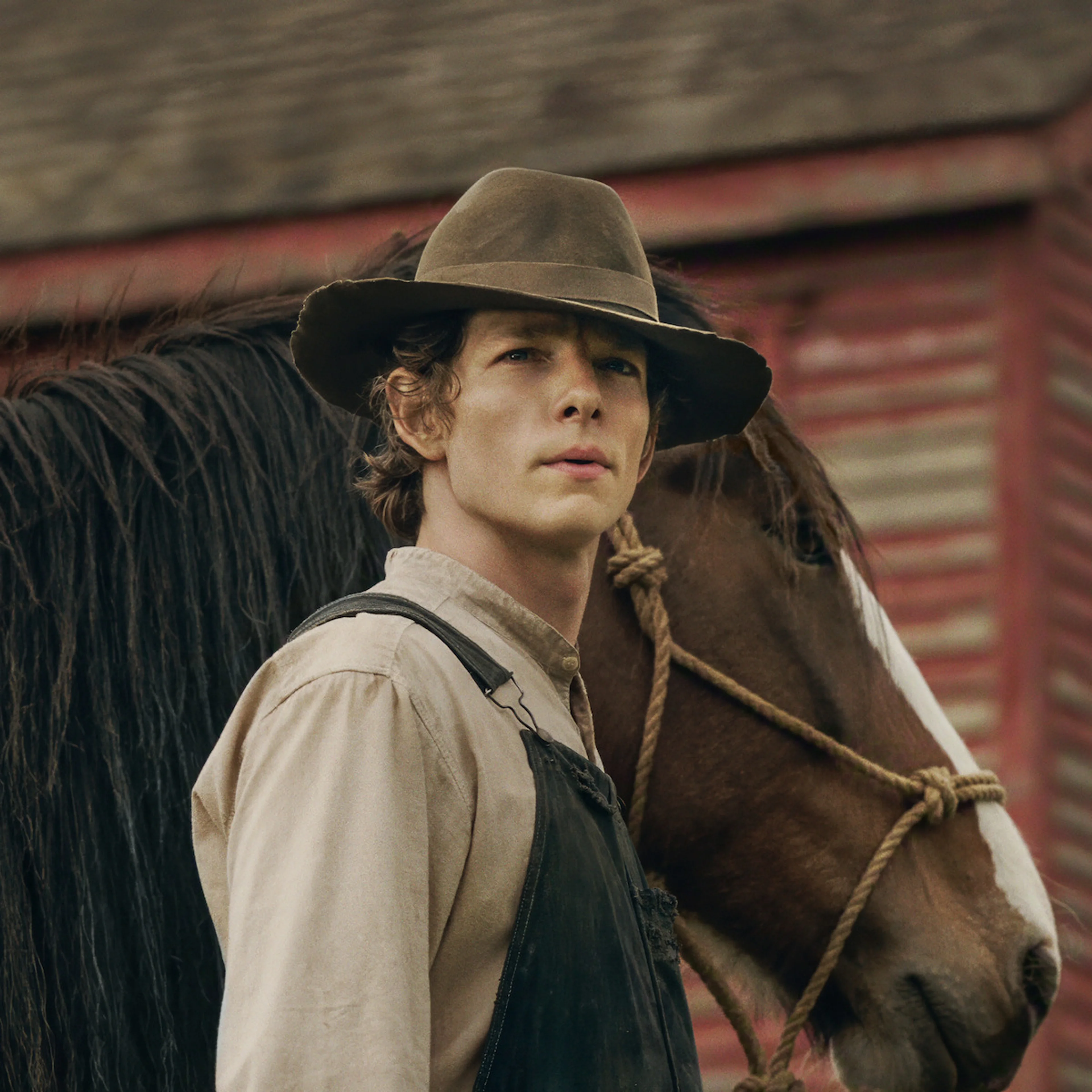 Mike Faist as Charles Trask wears a brown hat and overalls beside a horse.