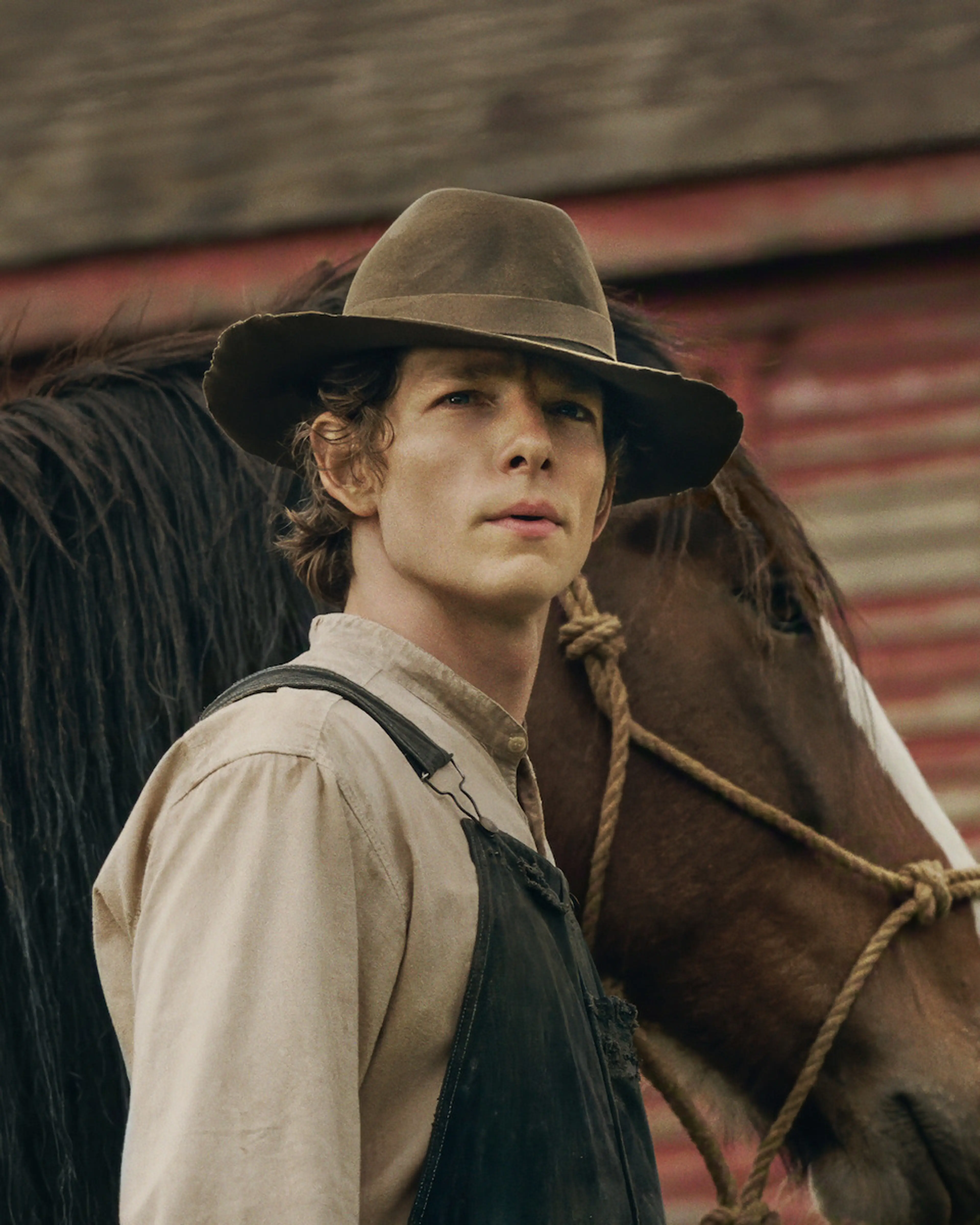Mike Faist as Charles Trask wears a brown hat and overalls beside a horse.