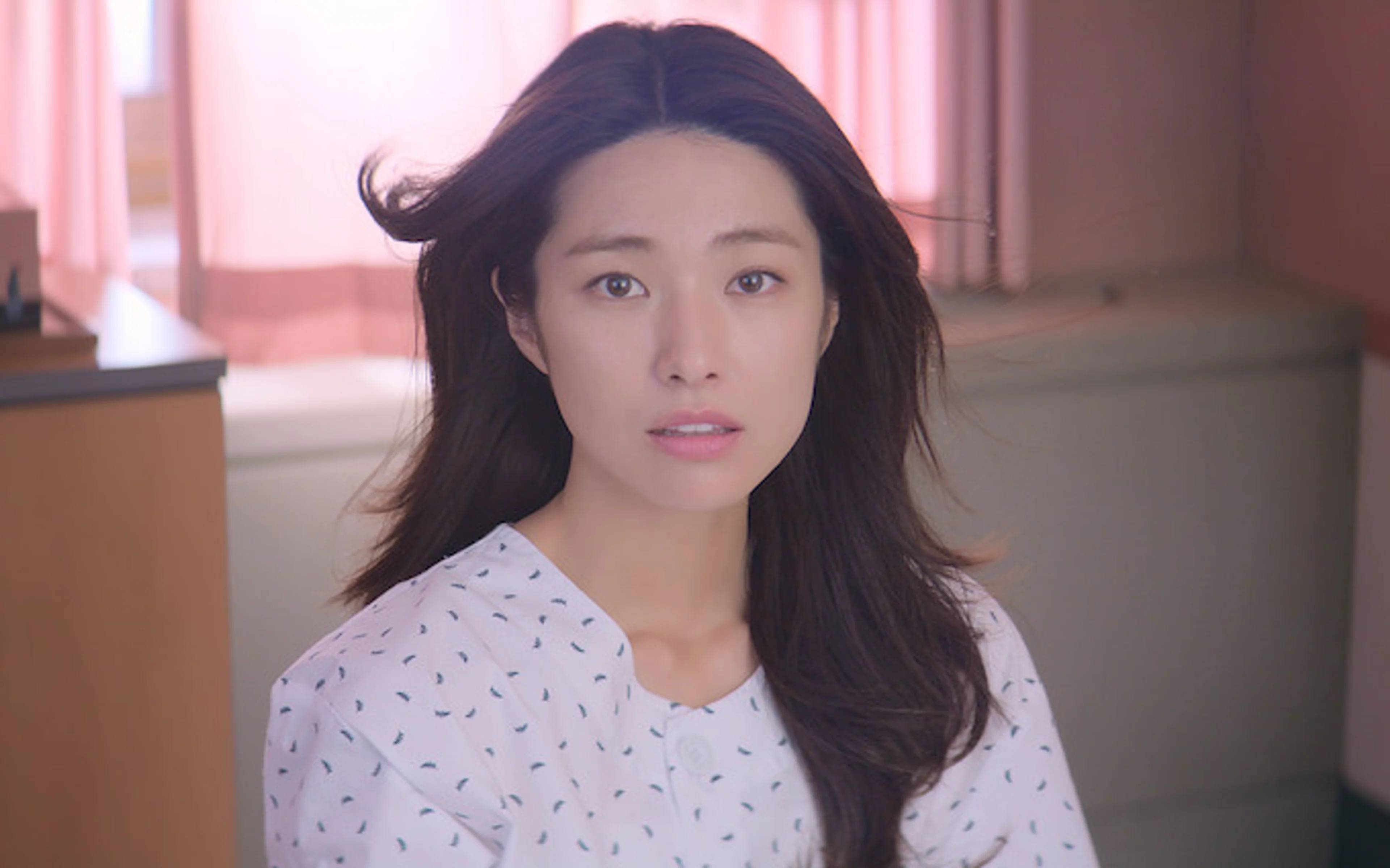 Ha Young faces forward in a patterned hospital gown in a bright hospital room.