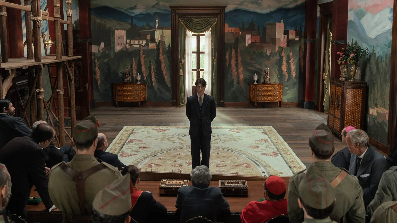 Carlos stands alone before seated officials in an ornate room.