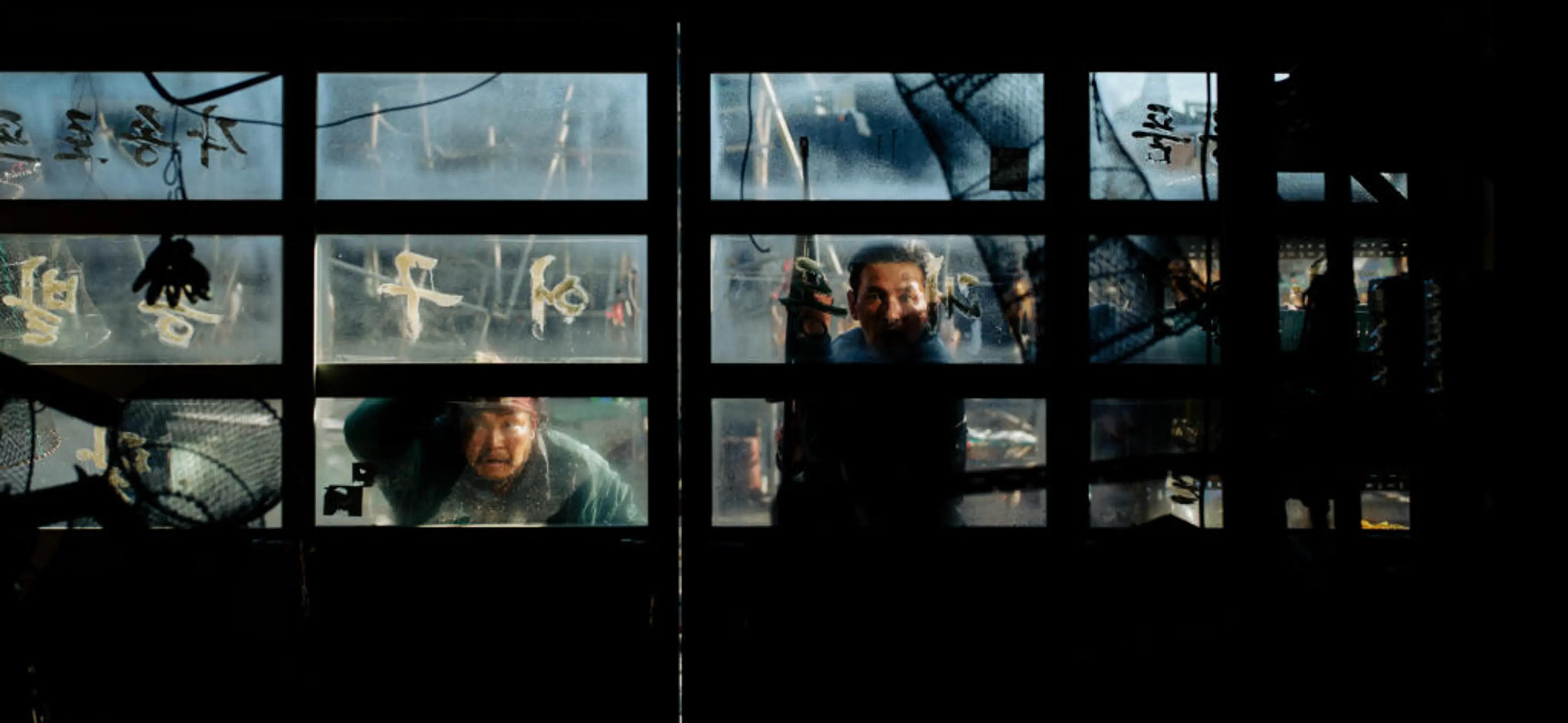 Two men look through a damaged window with writing on the glass.